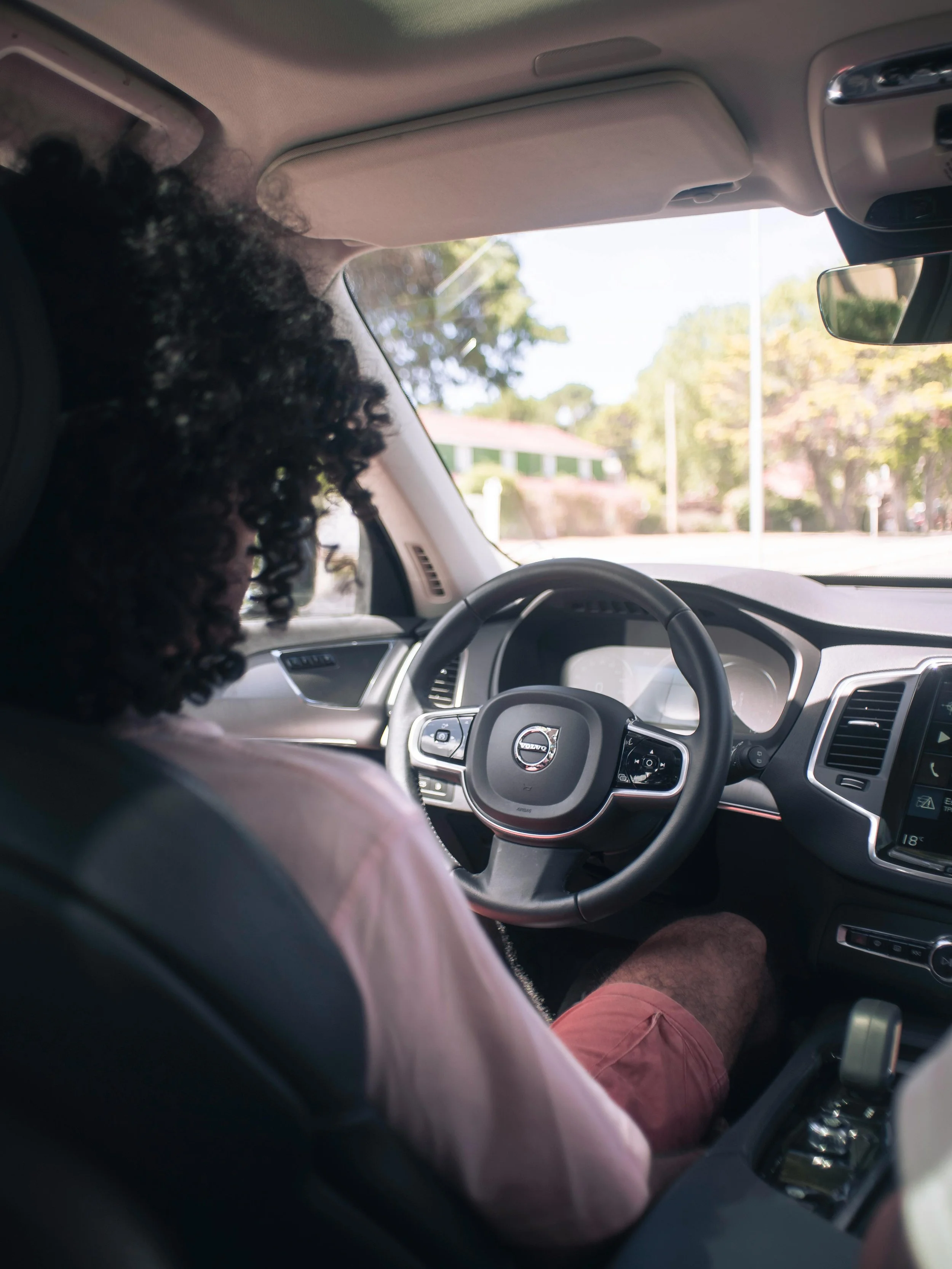 Person driving a Volvo car on a sunny day, with trees and houses visible outside.