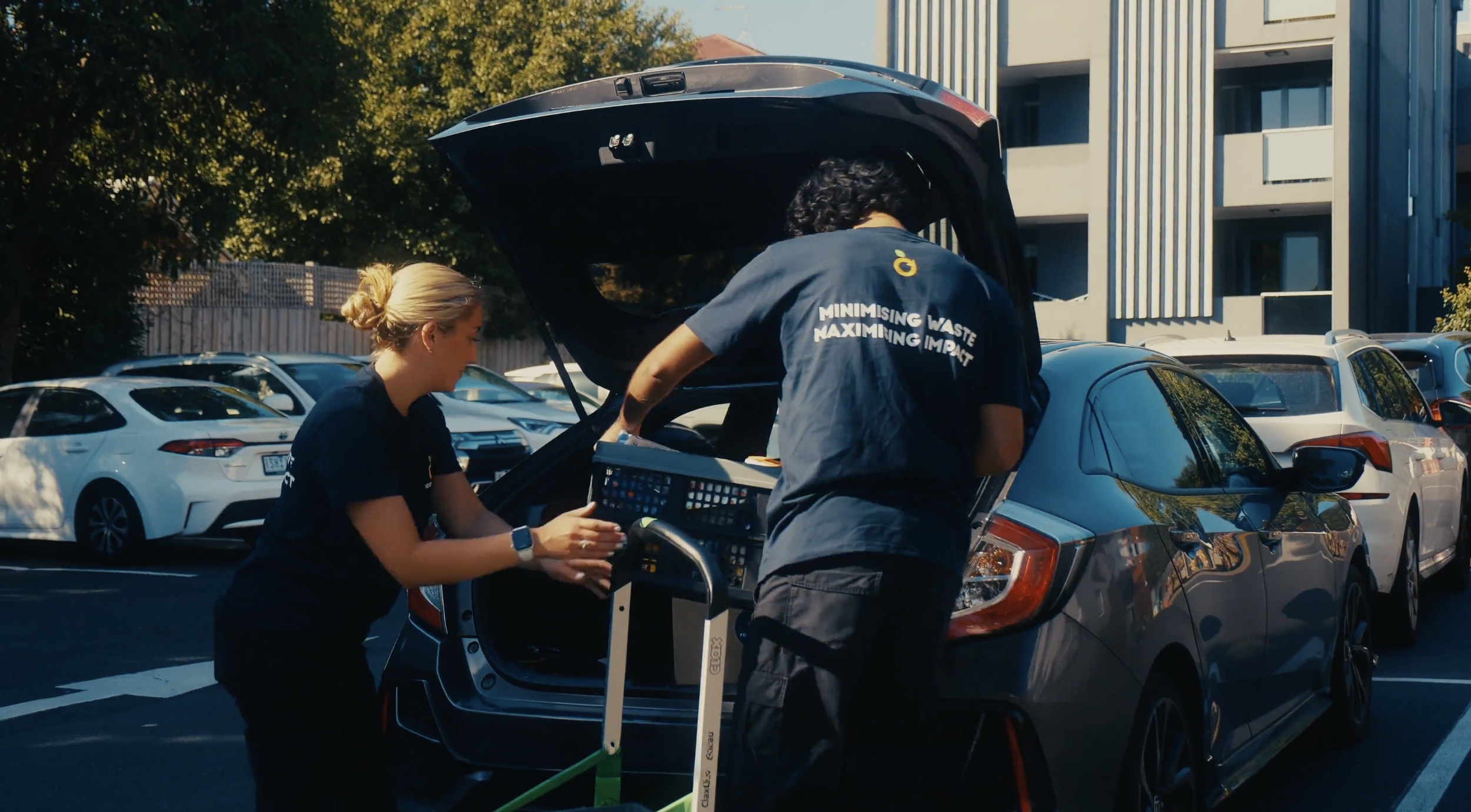 Two people loading items into the trunk of a silver car in a parking lot, with multiple parked cars and modern buildings in the background.