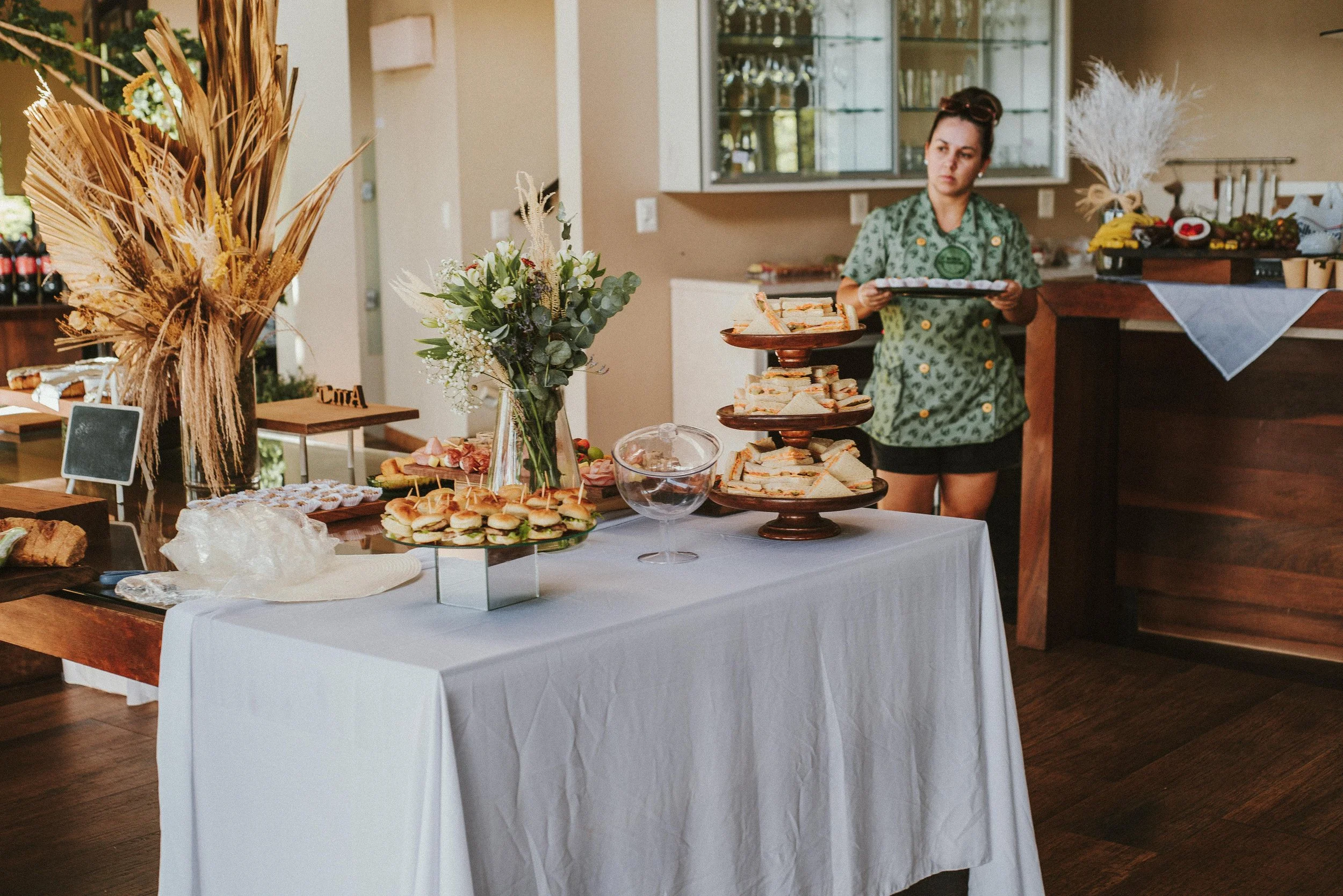 A buffet table covered with a white tablecloth, holding assorted finger foods including sandwiches, small burgers, and pastries. A woman in a green patterned apron stands behind the table holding a tray. Decor includes a large dried plant arrangement, a bouquet of flowers, and a glass bowl. The setting appears to be a catered event or gathering in a cozy indoor space with natural light.