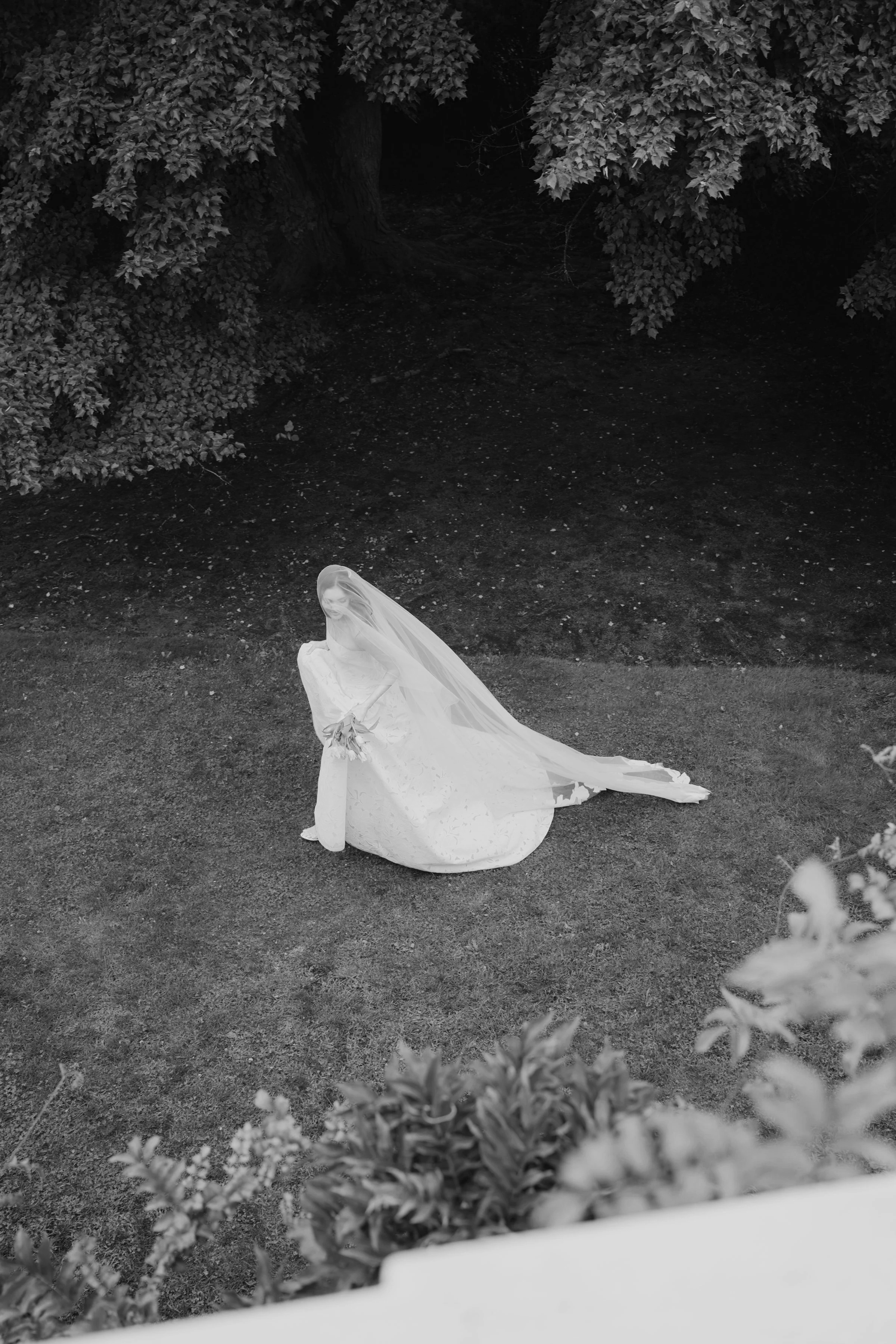 A bride in a wedding dress with a veil, sitting on the grass in a garden, holding a bouquet and facing toward the camera.