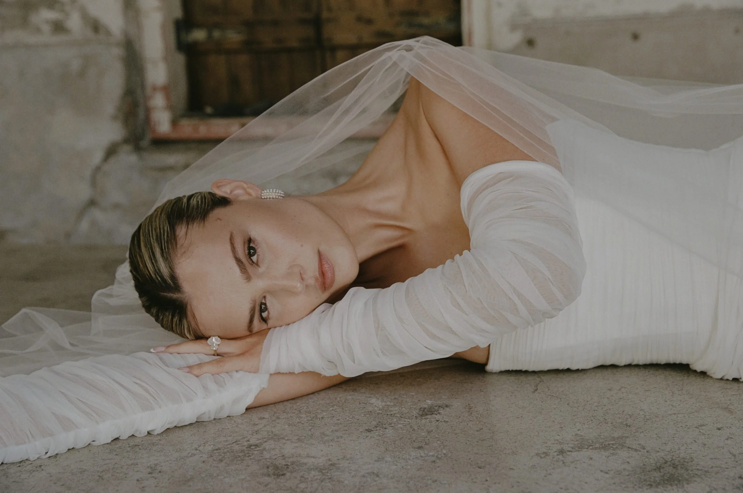A woman in a white dress with long sheer sleeves lying on a floor, resting her head on her arm, with a veil over her head, in an indoor setting with a rustic background.