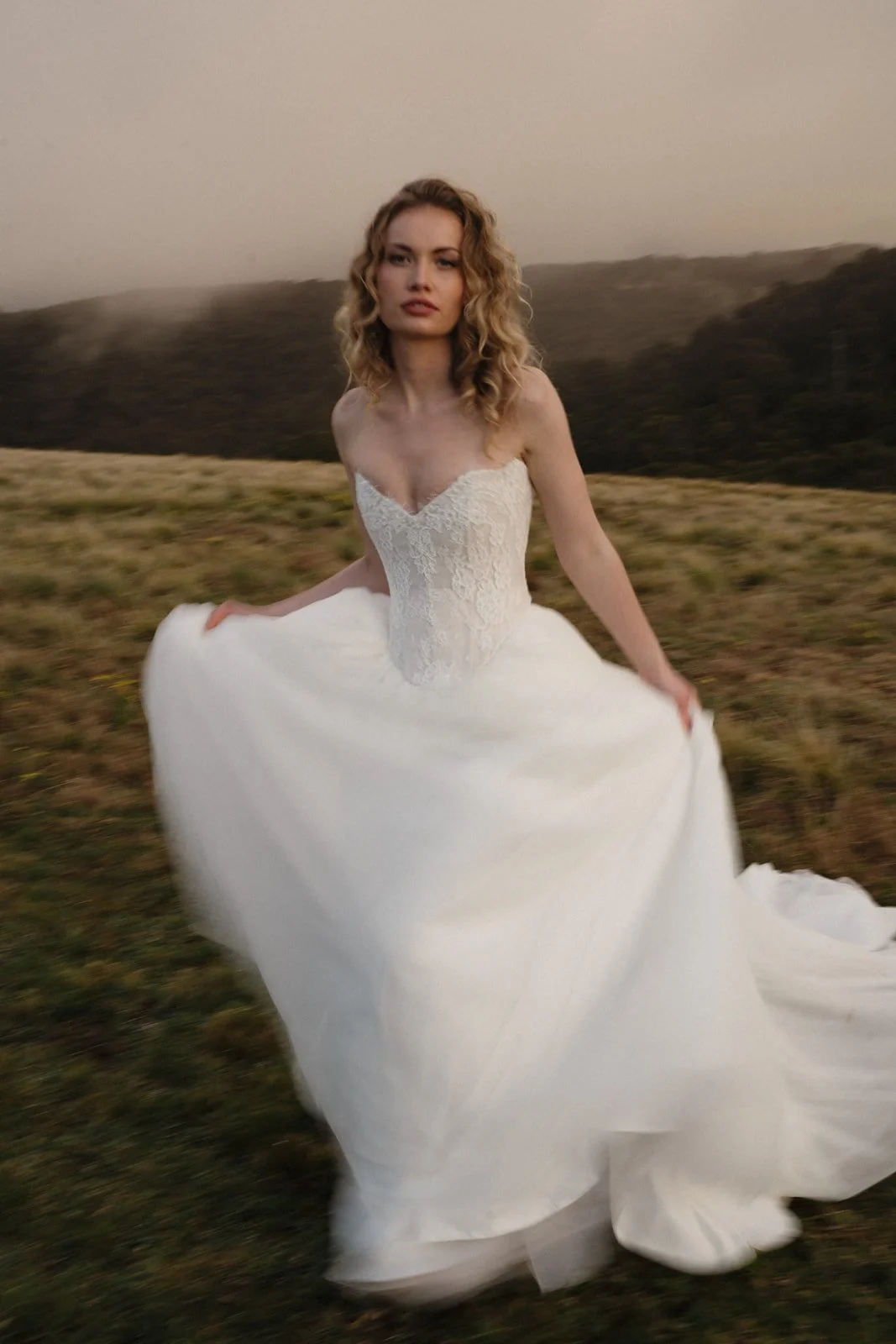 A woman in a white wedding dress standing in a grassy field with hills and trees in the background.