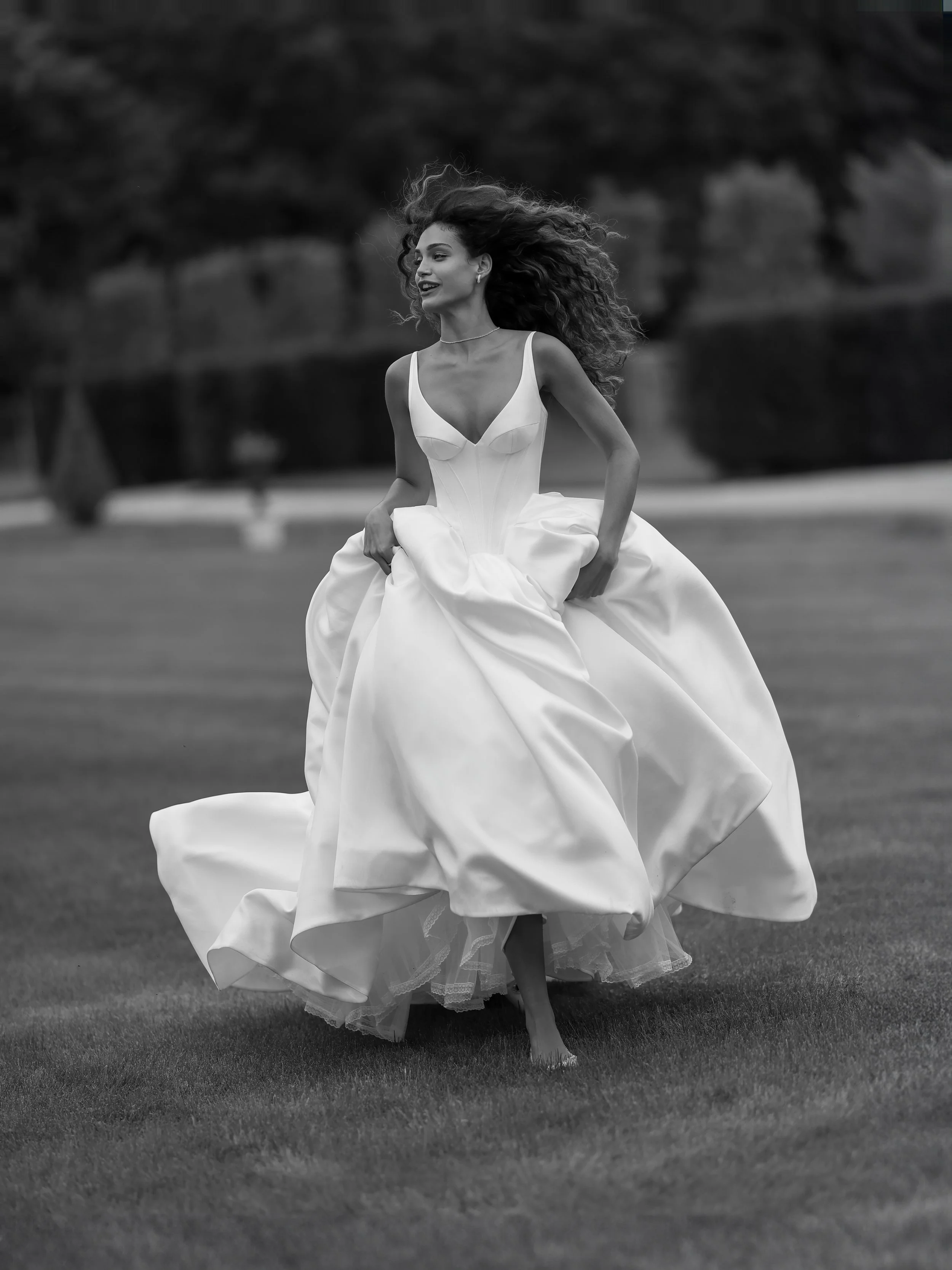 A woman in a white wedding dress running barefoot on grass, with trees in the background.