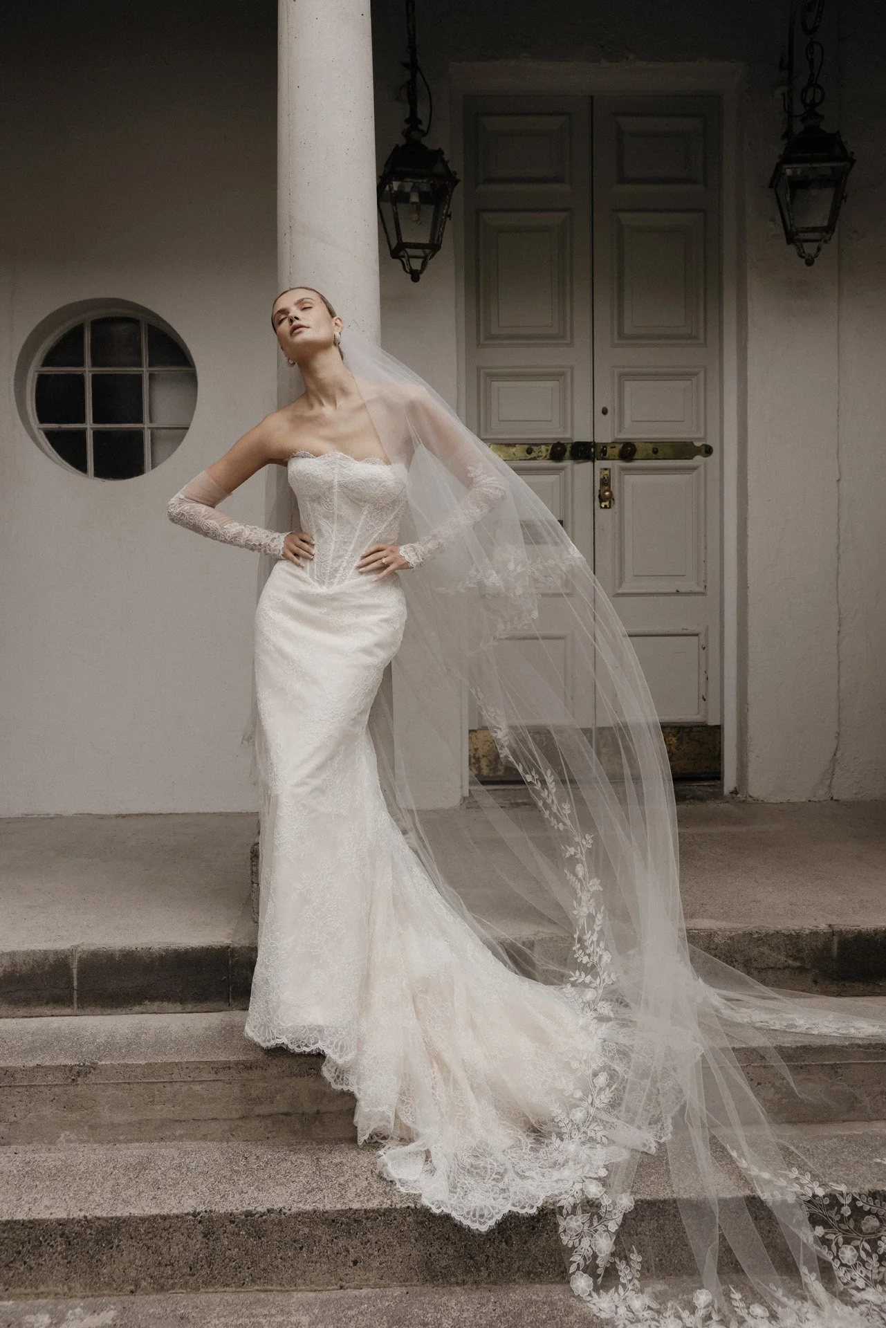 Bride in a white wedding gown with lace details, long veil, standing on steps in front of a white door with lanterns on either side.