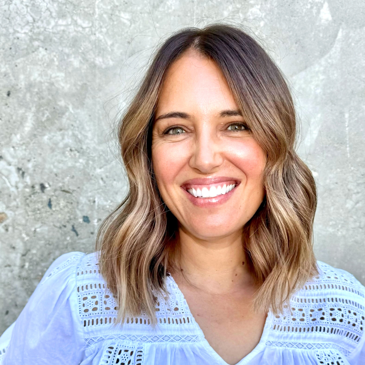 A smiling woman with shoulder-length wavy brown hair and light skin, wearing a light blue embroidered top, standing in front of a textured gray wall.