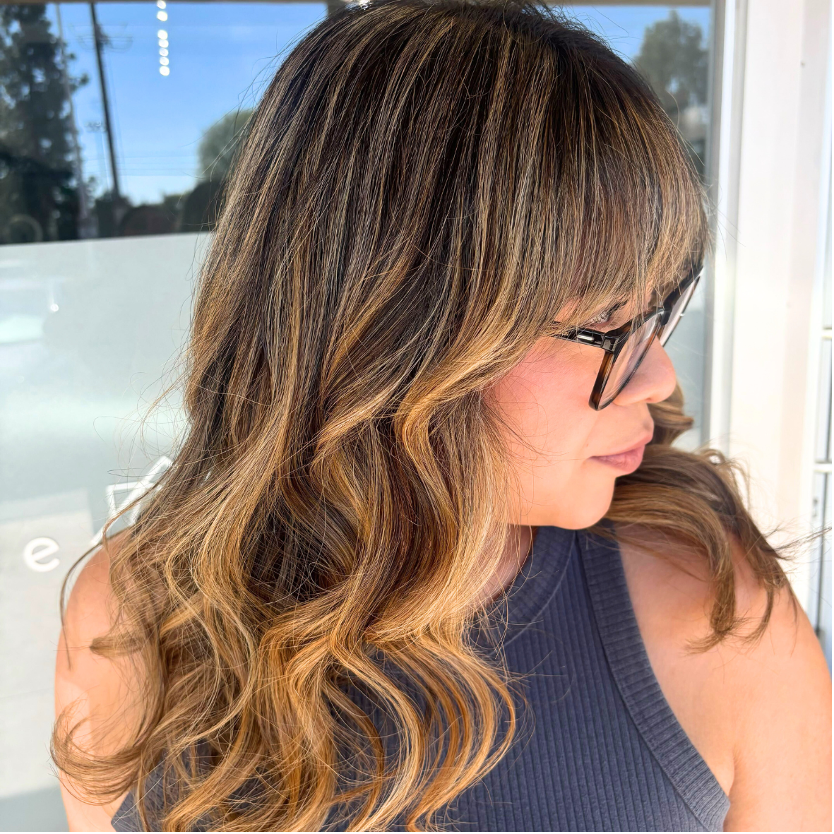 Close-up of a woman with wavy brown hair with blonde highlights, wearing glasses, looking to the side inside a bright room.