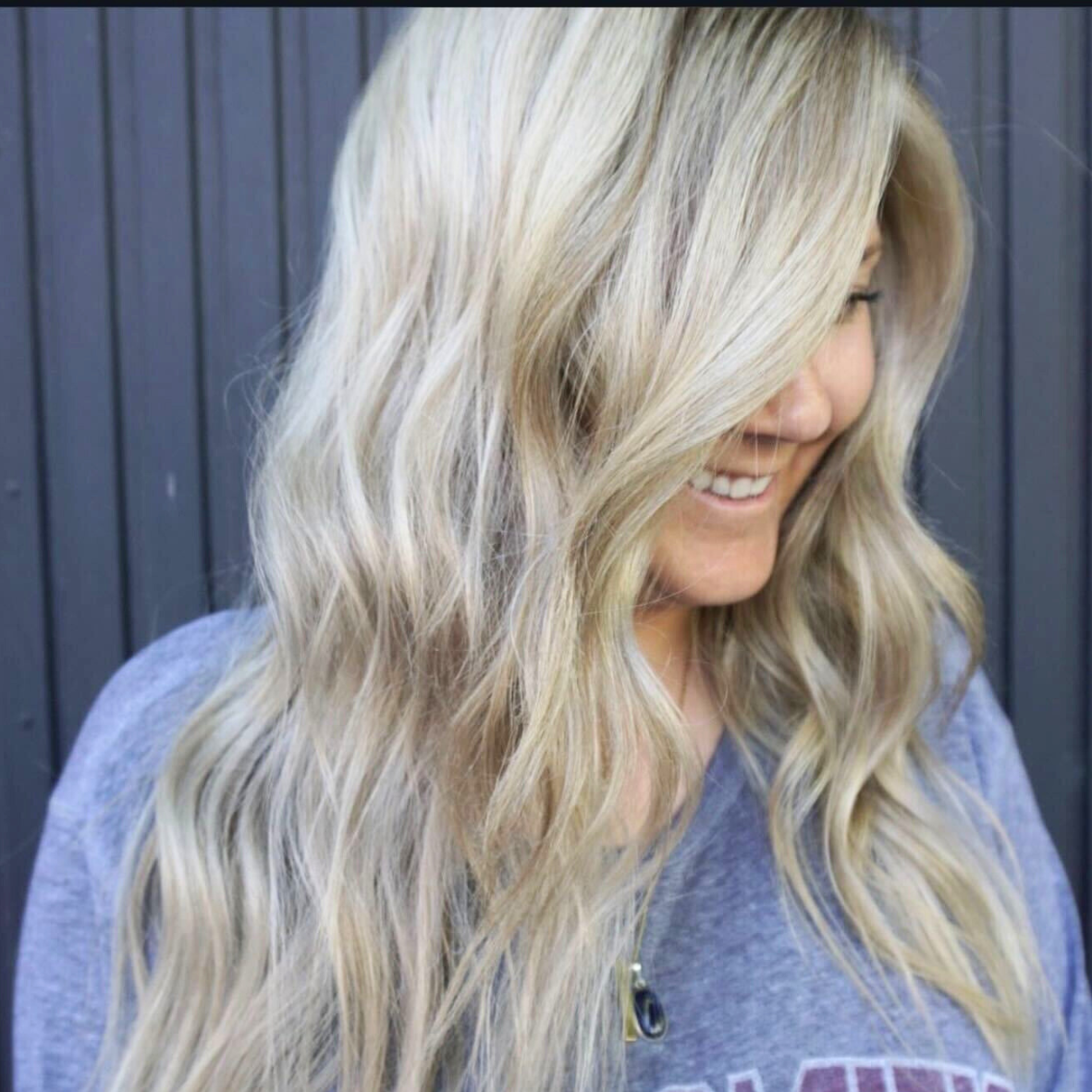 Woman with long, wavy blonde hair smiling and looking downward, standing in front of a dark wooden wall.