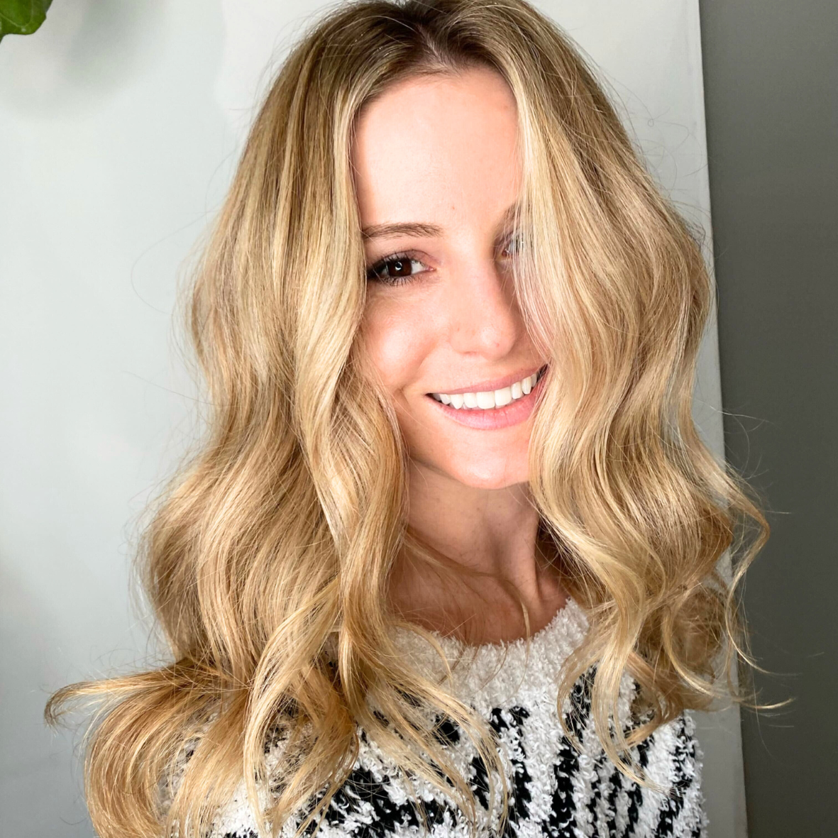 A woman with long, wavy blonde hair smiling at the camera, wearing a black and white patterned top.