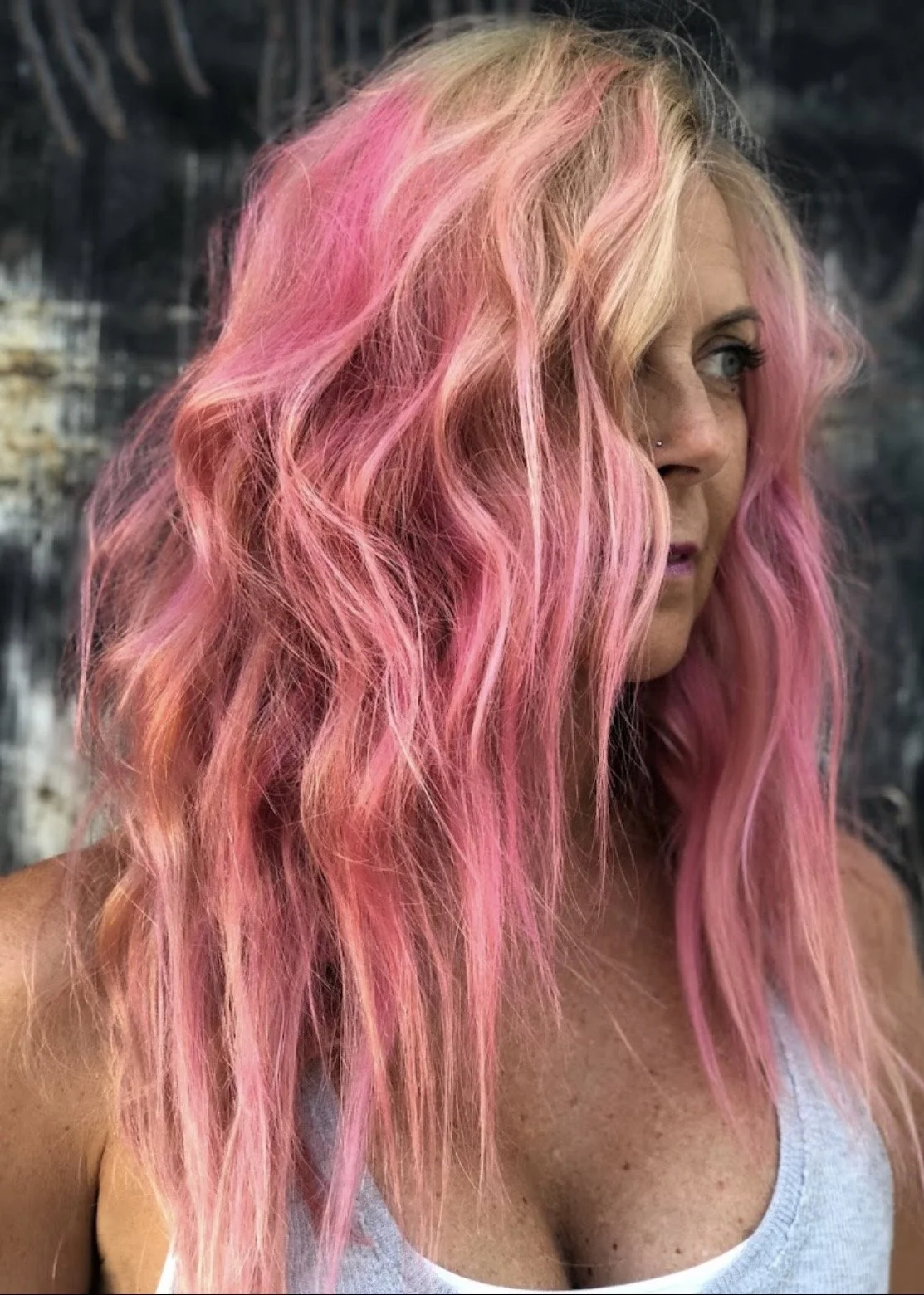 A woman with long, wavy pink hair and blonde roots, wearing a light gray tank top, looking to the right, with a dark, textured background.