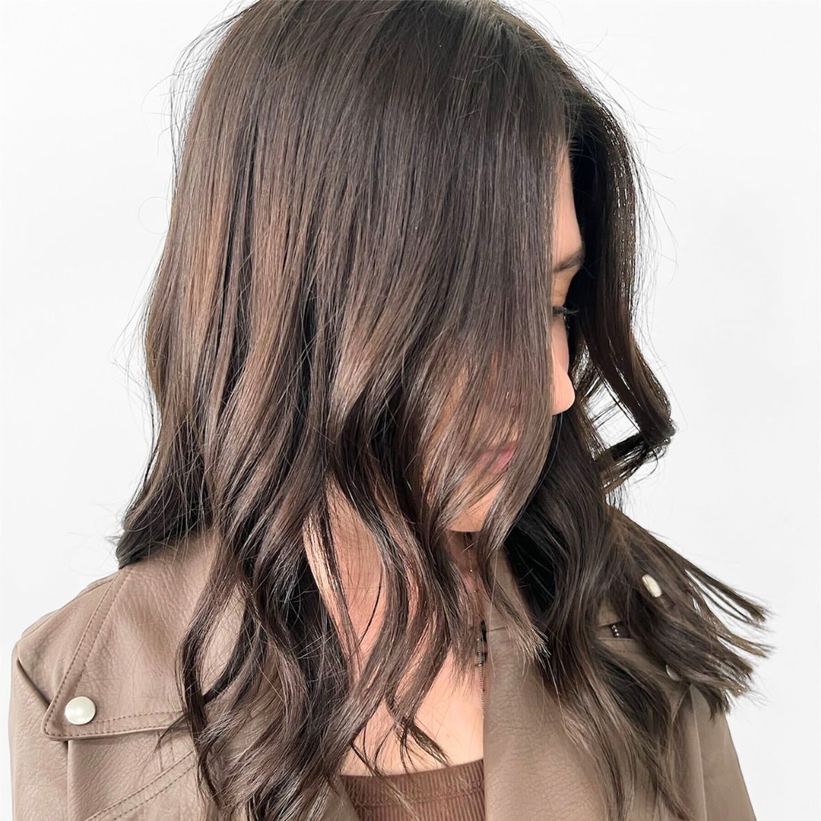 Side view of a woman with shoulder-length wavy brown hair with highlights, wearing a beige jacket with pearl buttons.