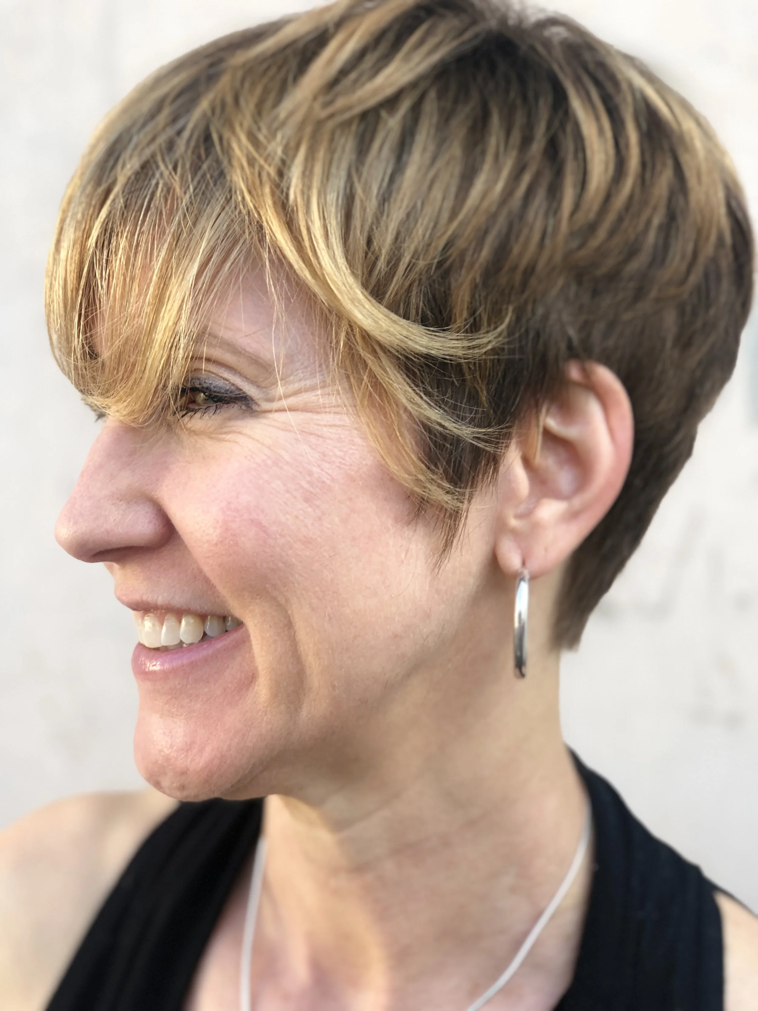 A smiling woman with short, layered blonde hair and a tan complexion, wearing hoop earrings, a black top, and white earphones, against a light background.