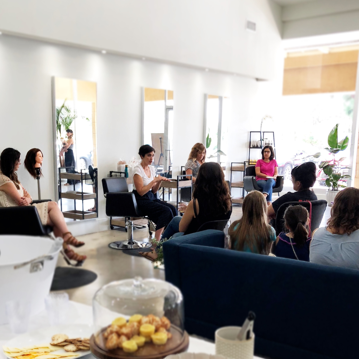 A group of women and children attending a workshop or seminar in a bright, modern space with large mirrors and green plants. A woman is speaking to the group while others listen, some taking notes.