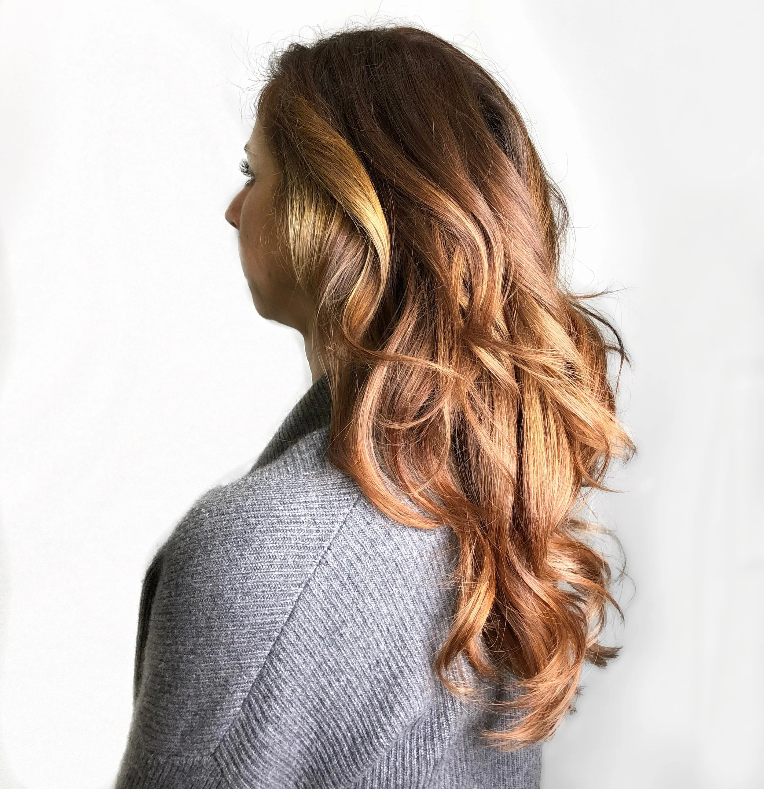 Profile of a woman with wavy, reddish-brown hair, wearing a gray sweater, against a plain white background.