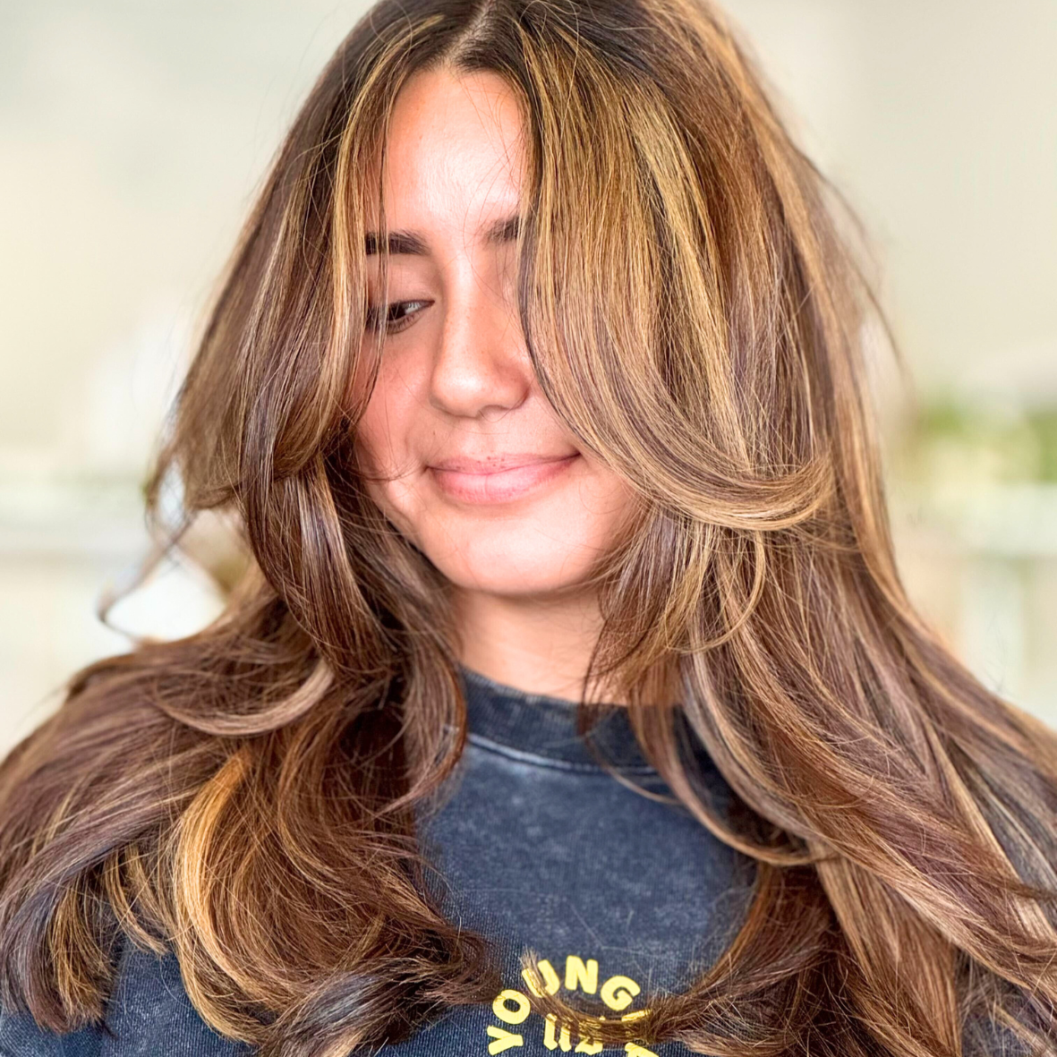A woman with long, wavy brunette hair with highlights, smiling softly, wearing a dark gray t-shirt.