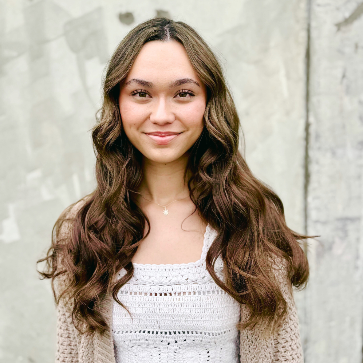 A young woman with long, wavy brown hair, smiling, standing outdoors against a light gray concrete wall, wearing a white crocheted top and a delicate necklace.