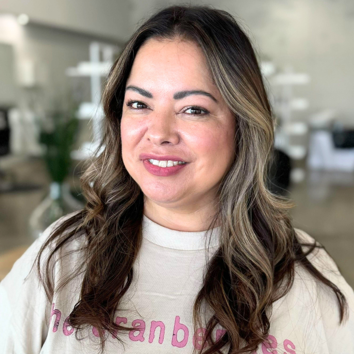 A woman with wavy brown hair and light skin smiling at the camera, wearing a beige top with pink lettering.