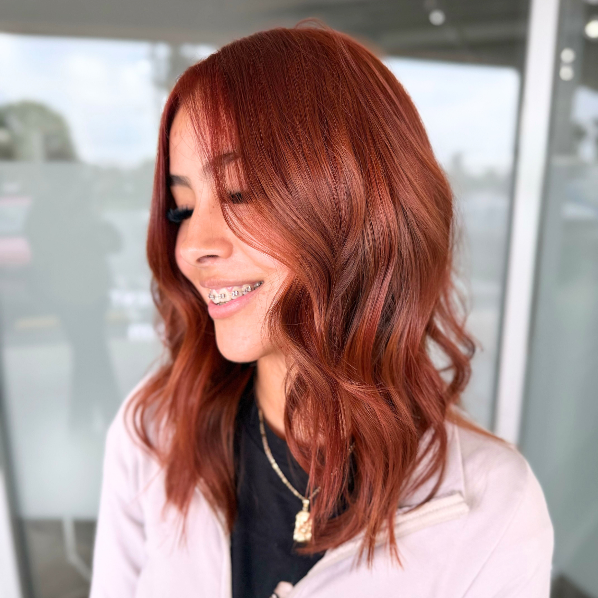 Profile of a woman with wavy red hair, smiling with braces, wearing a black top, a light-colored jacket, and layered necklaces.