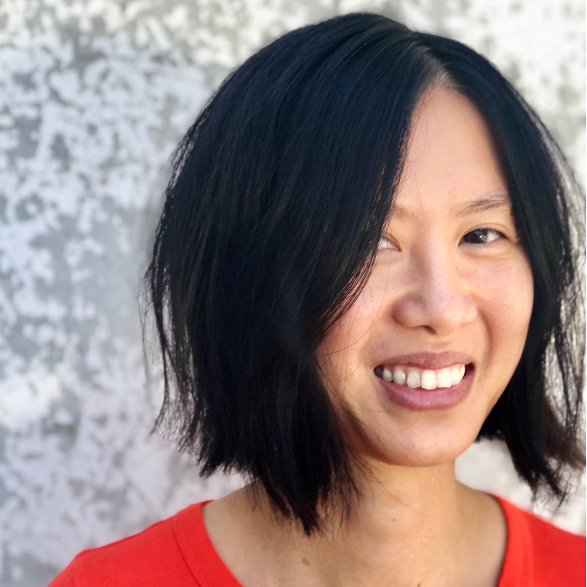 Close-up of a woman with black hair and a red shirt smiling against a light patterned background.