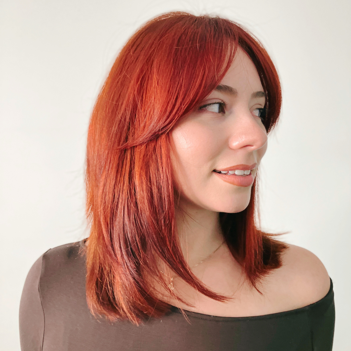 A woman with shoulder-length red hair wearing a black off-the-shoulder top, smiling slightly and looking to the side against a plain white background.