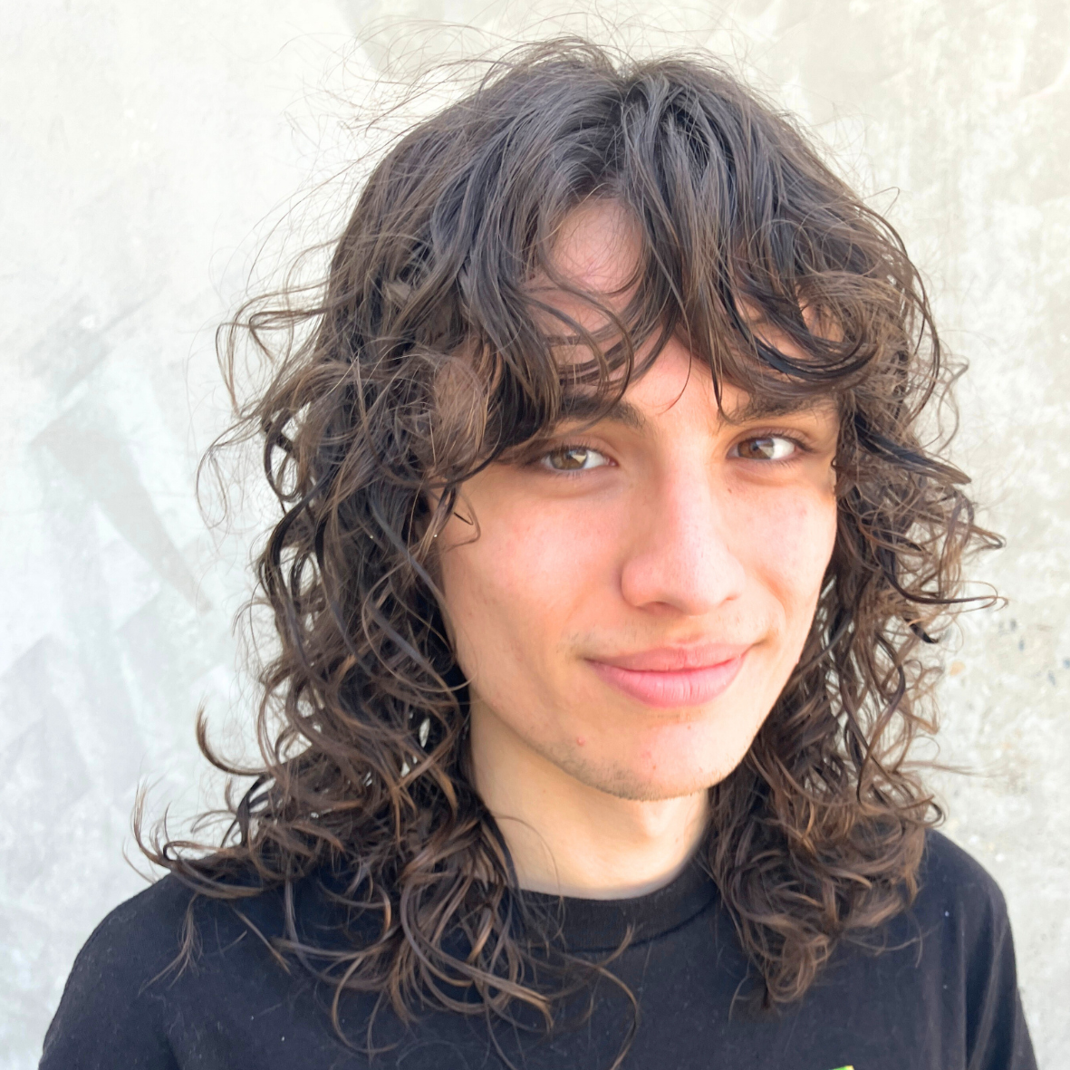 A young person with curly brown hair, light skin, and brown eyes, smiling slightly, wearing a black shirt, standing against a light-colored textured wall.