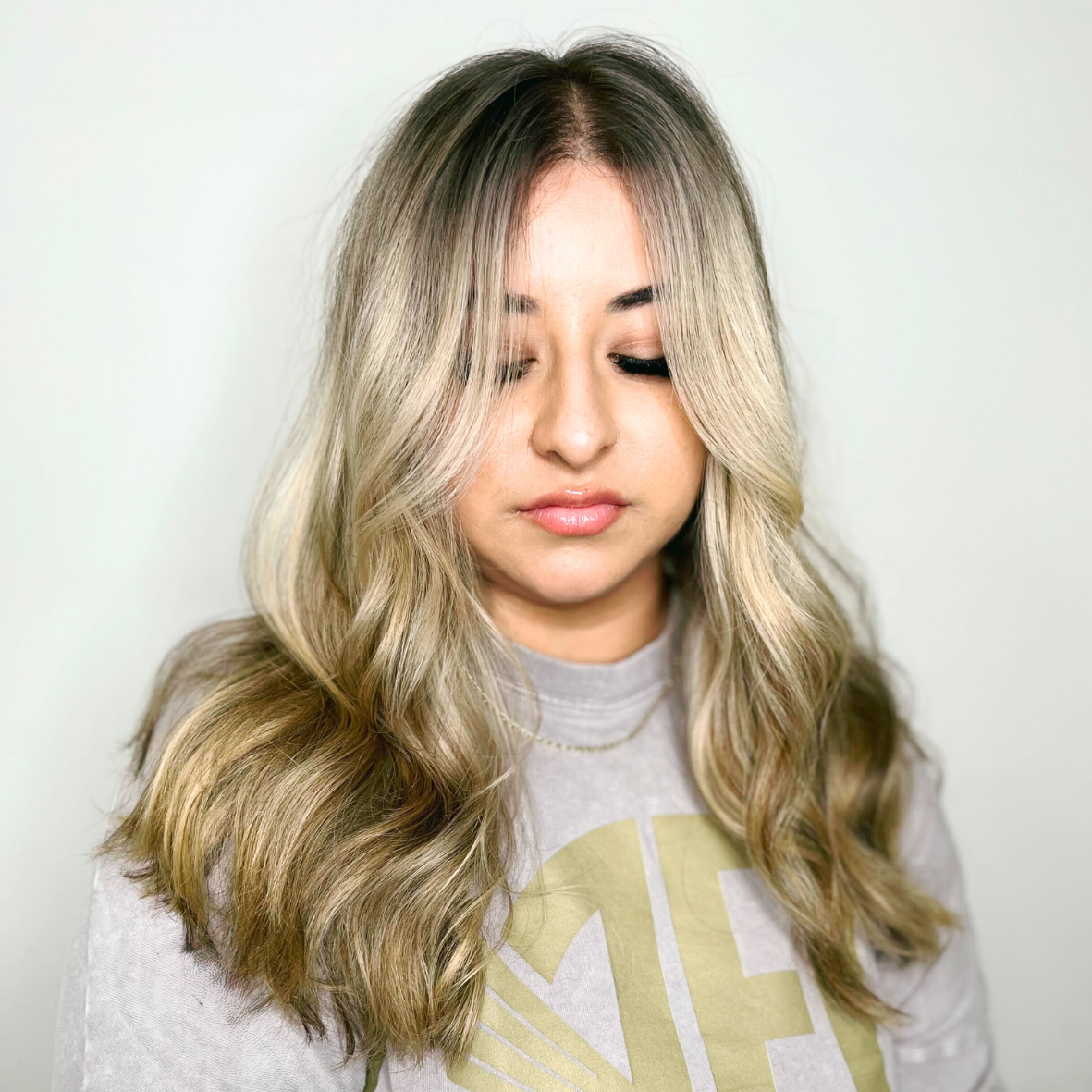 A woman with long, wavy blonde hair, wearing a light gray shirt with a yellow graphic, looking downward with a neutral expression.
