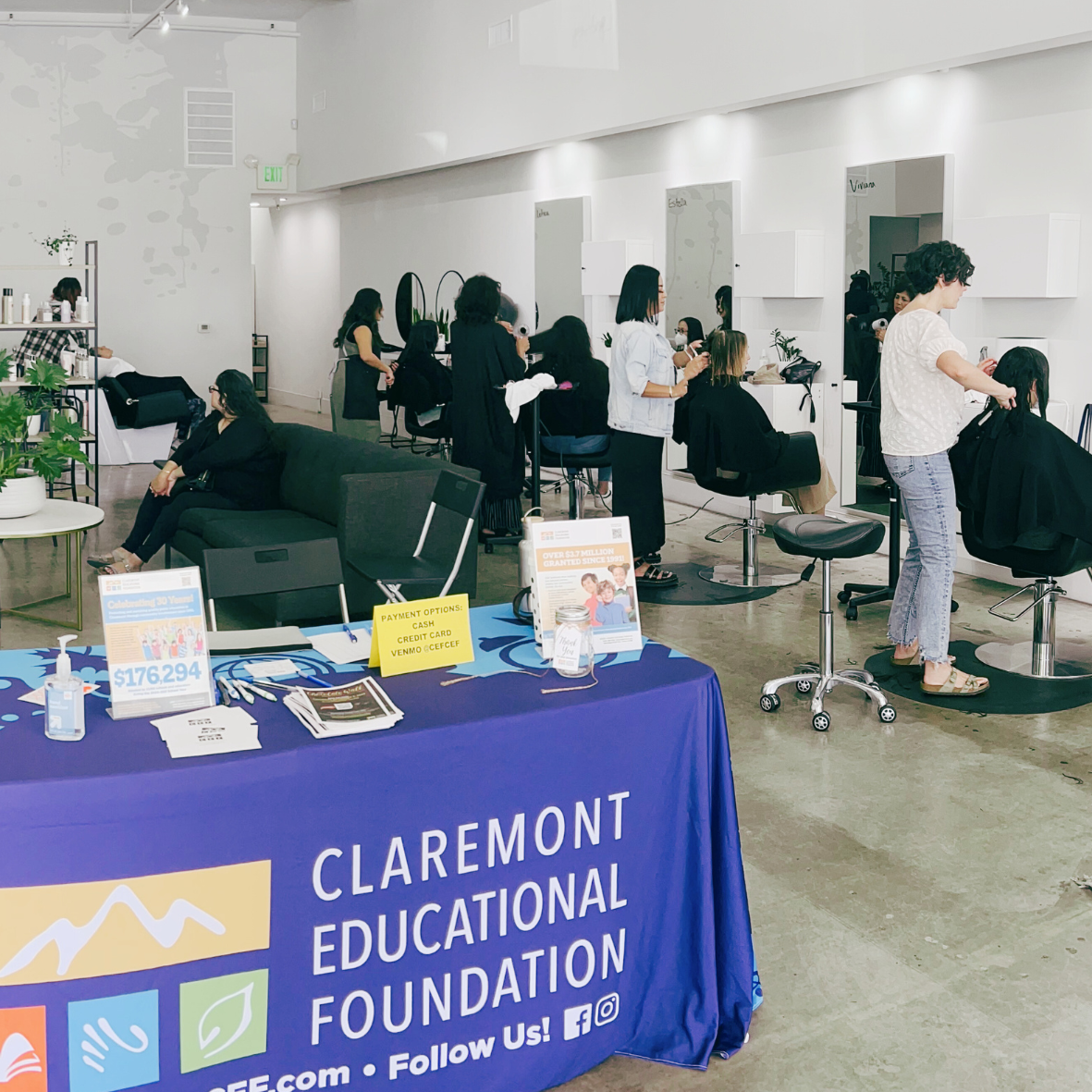 Interior of a hair salon or beauty studio with multiple people getting hair treatments, and a table at the front with informational materials and signs, including a blue tablecloth with the Claremont Educational Foundation logo.