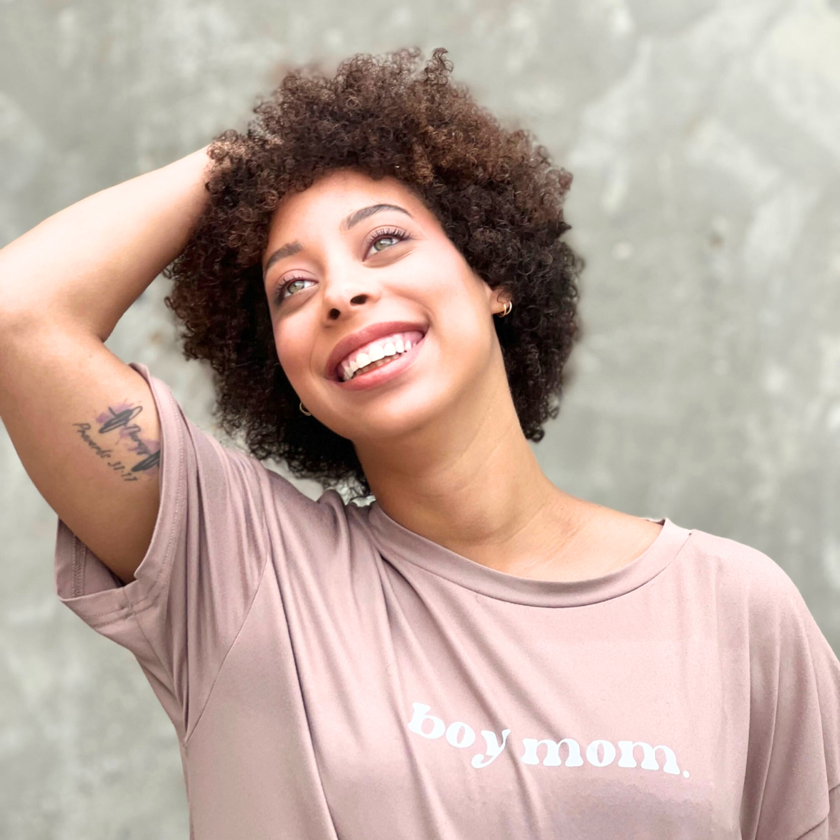 A woman with curly hair smiling, wearing a beige T-shirt that says 'boy mom,' standing against a gray concrete background.