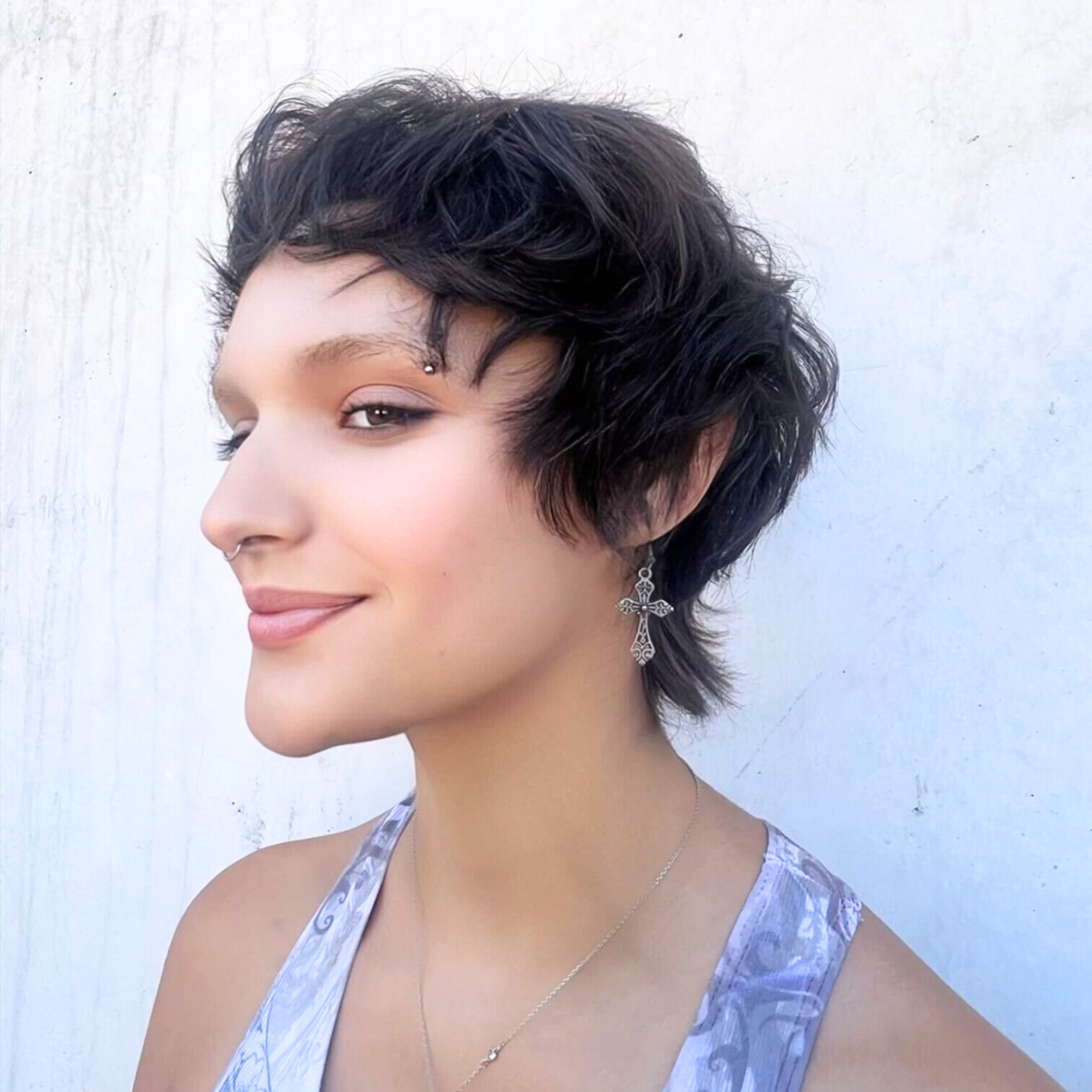 A woman with short dark curly hair, wearing earrings and a light-colored top, smiling and looking to her left, standing against a white background.