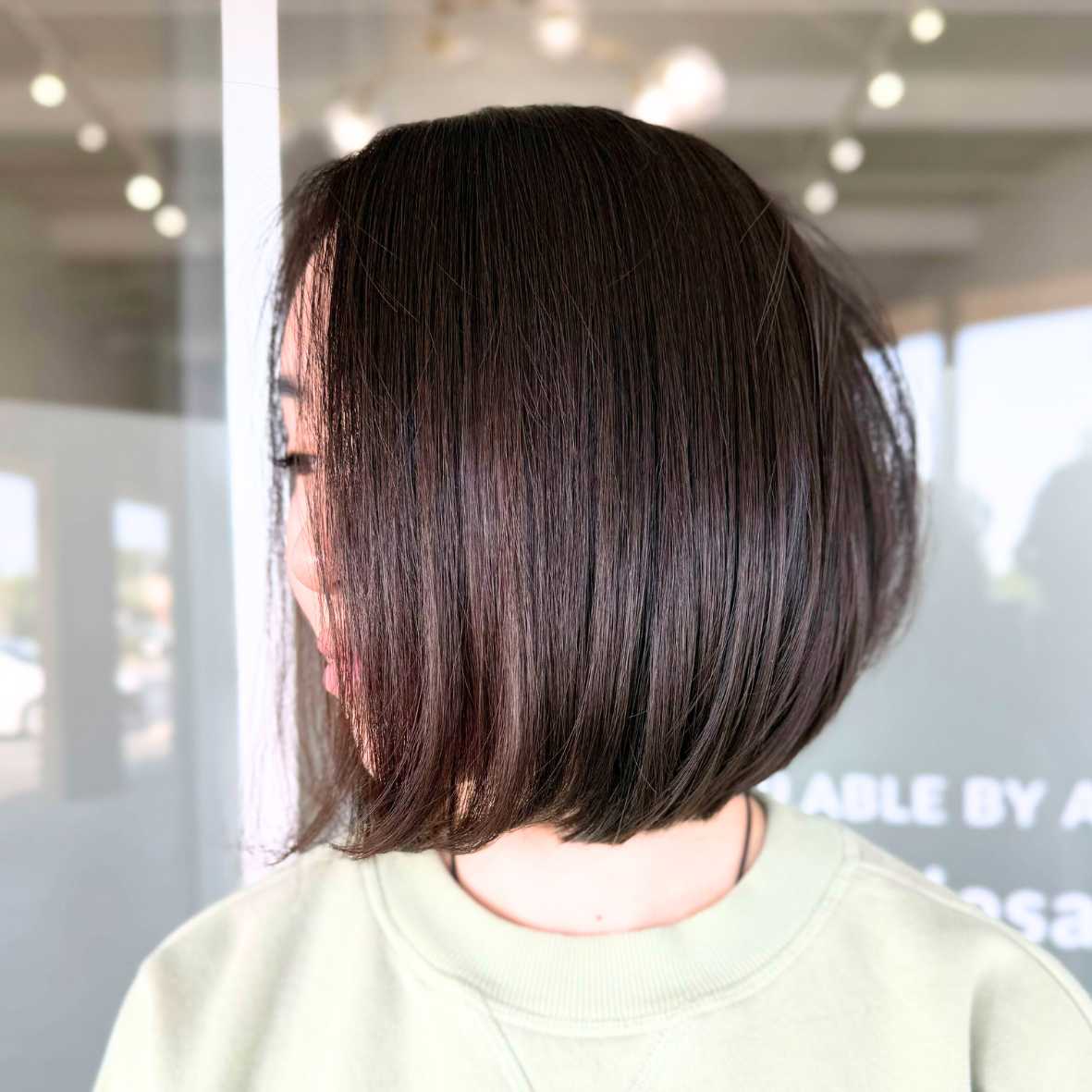 A person with a sleek, straight, shoulder-length bob haircut in dark brown, standing indoors near a glass window with natural light.