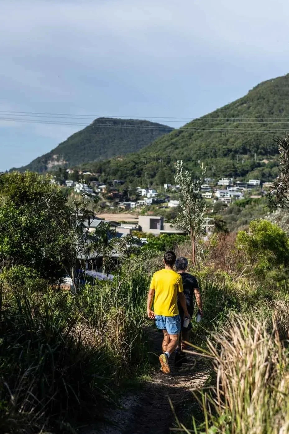 Two people hiking on a narrow dirt trail surrounded by green bushes and trees with a town and mountains in the background.