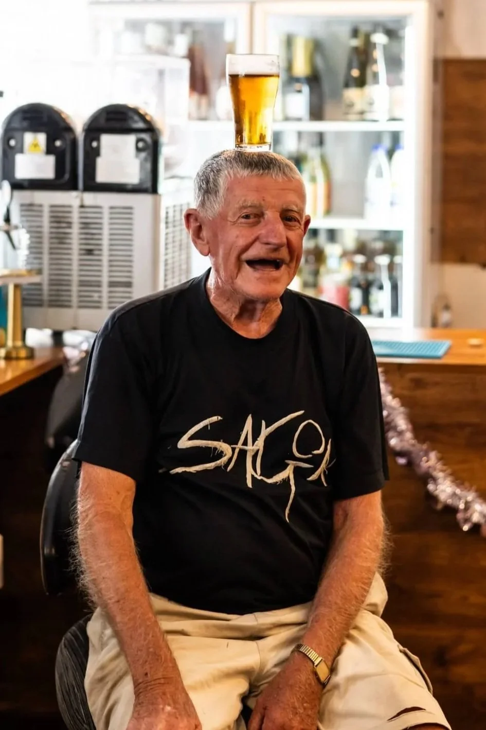 An elderly man sitting in a restaurant with a glass of beer balanced on his head, smiling and wearing a black T-shirt with the word 'SAGA' on it.