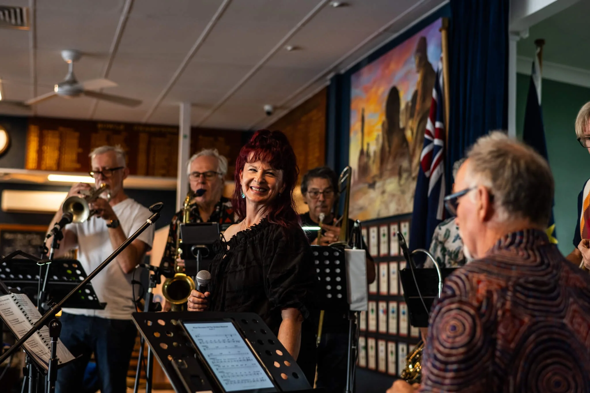A smiling woman holding a microphone in front of a jazz band performing in a room decorated with flags and artwork.