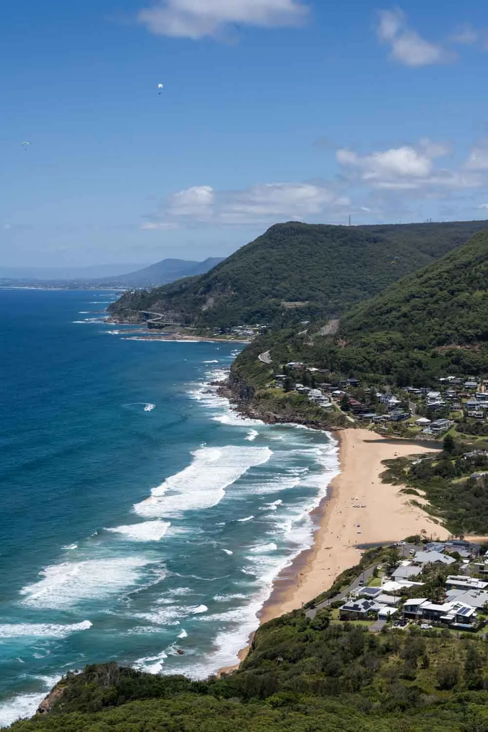 Aerial view of a coastline with a sandy beach, blue ocean waves, green hills, and residential houses along the shoreline, under a partly cloudy sky.