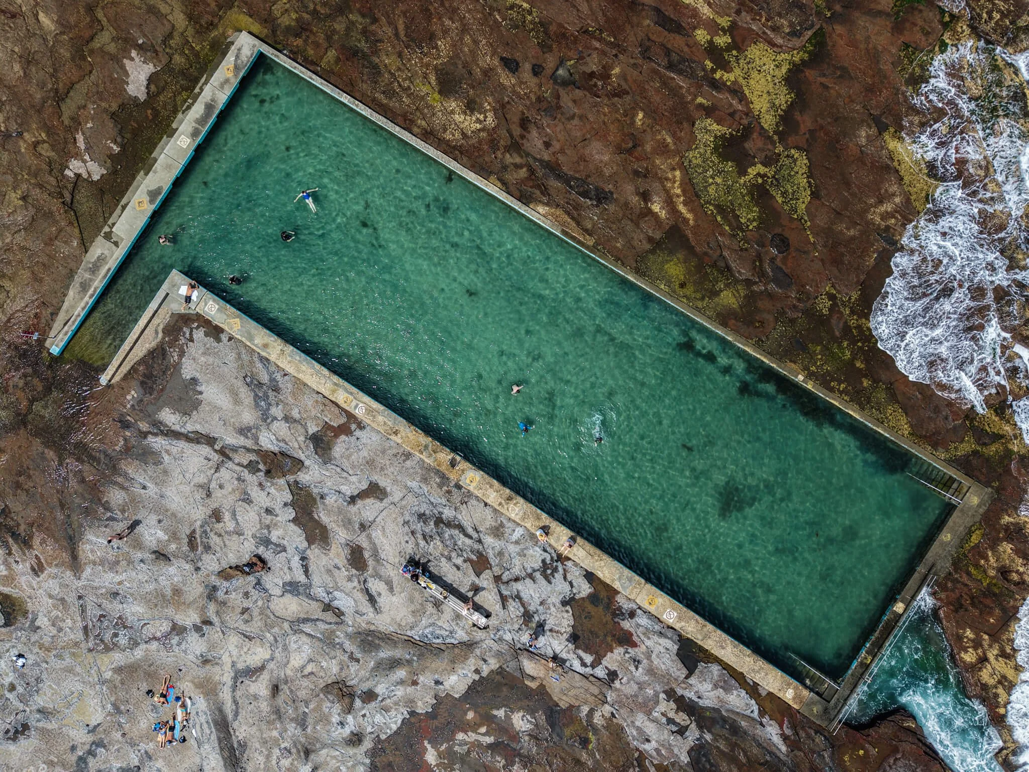 An aerial view of a rectangular swimming pool situated next to rocky terrain with waves crashing on the rocks at one end of the pool. Several people are in the pool, some swimming and others floating, with a few near the edge.