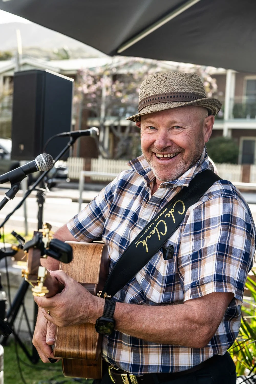 A smiling man wearing a straw hat and plaid shirt playing an acoustic guitar outdoors with a microphone and speakers nearby, in a suburban neighborhood.
