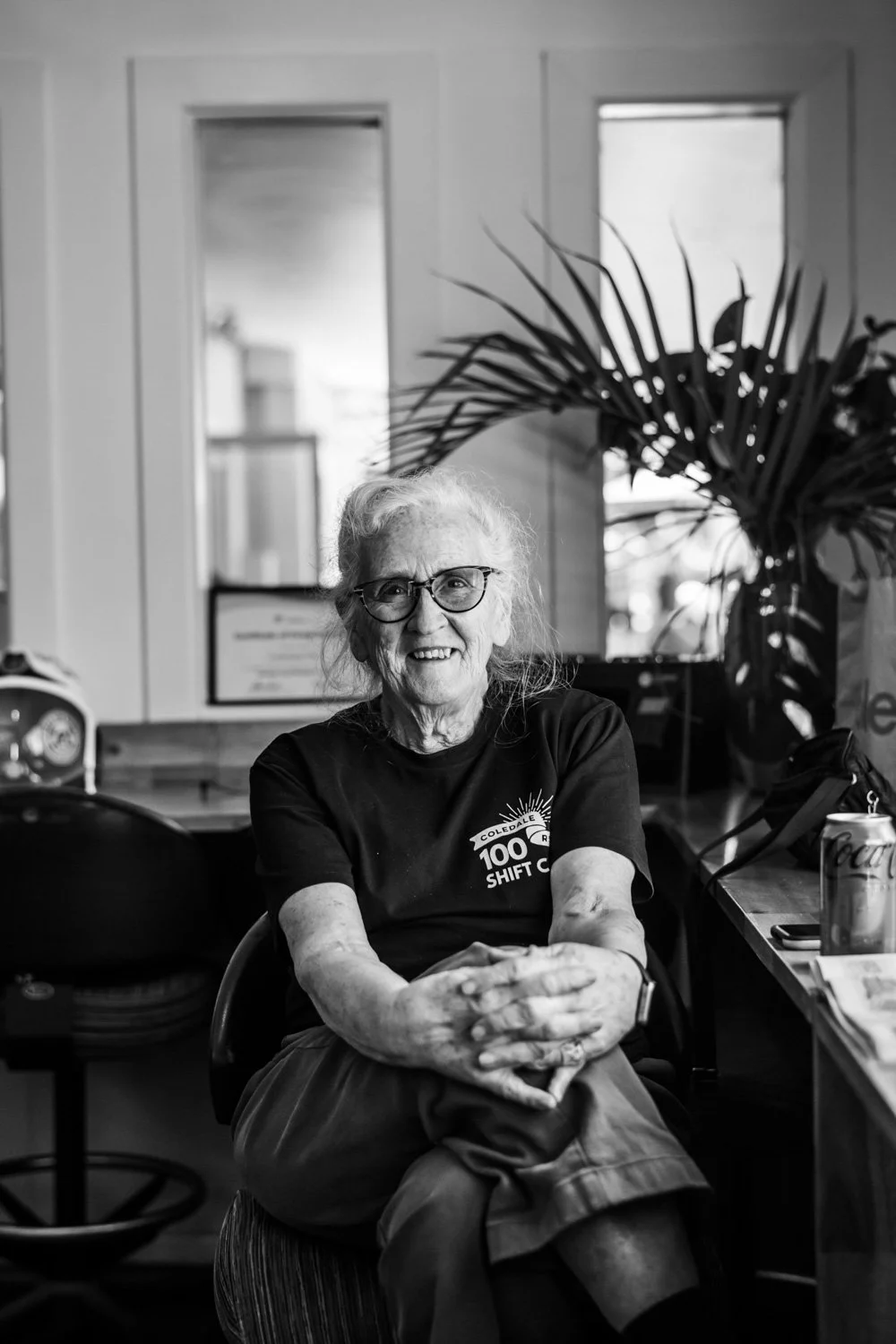 Older woman with glasses smiling, sitting at a table in a well-lit room with a large plant behind her.