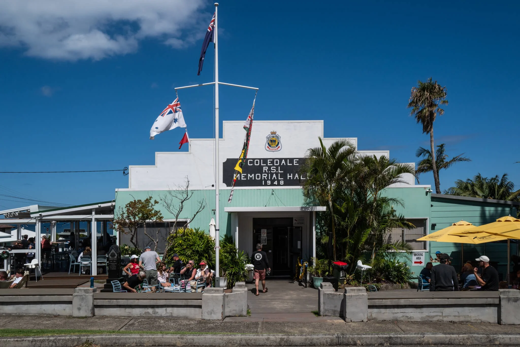 The Coedale RSL Memorial Hall, built in 1948, in Coedale with a white facade, surrounded by palm trees, people sitting outside at tables with yellow umbrellas, and a flagpole with the Australian flag and other flags, under a blue sky.