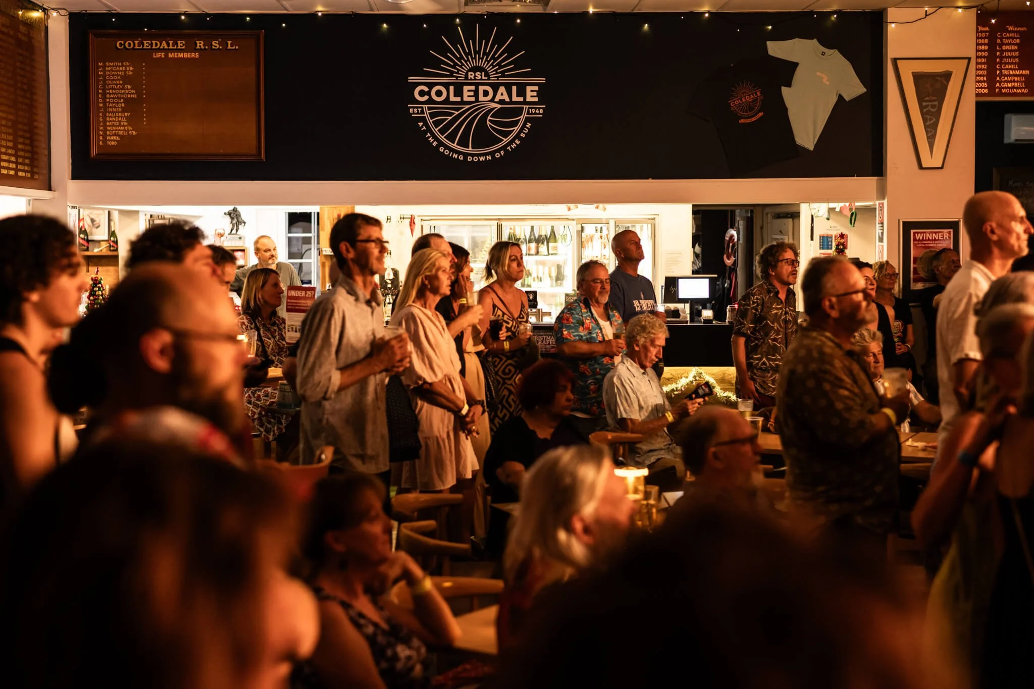 People gathered inside a bar or pub, watching a performance or event, with some standing and others sitting, illuminated by warm lighting, featuring a sign for Coledale R.S.L. and various decorations on the back wall.