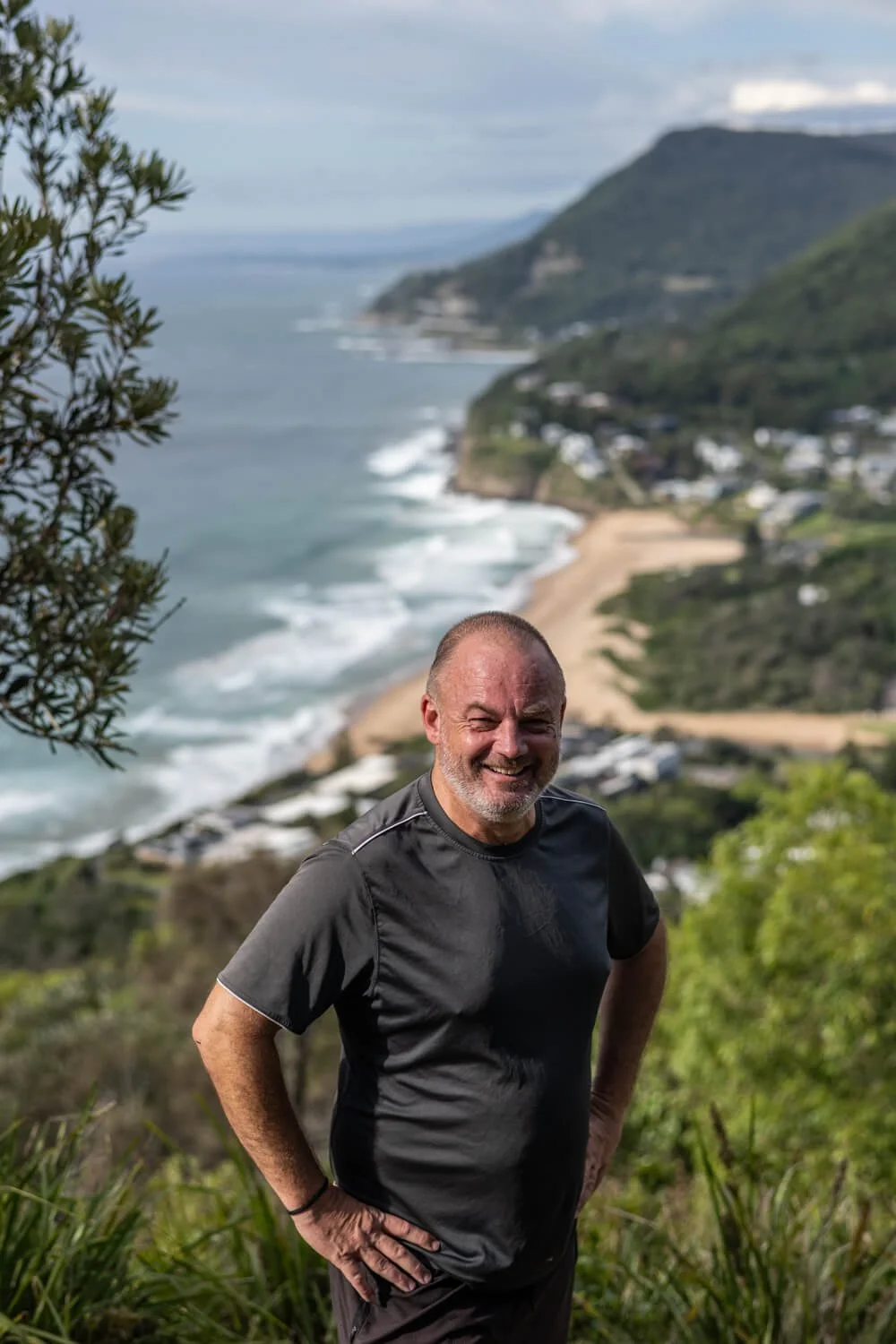 A smiling man standing outdoors on a hillside overlooking the coast, with trees and a beach visible in the background.