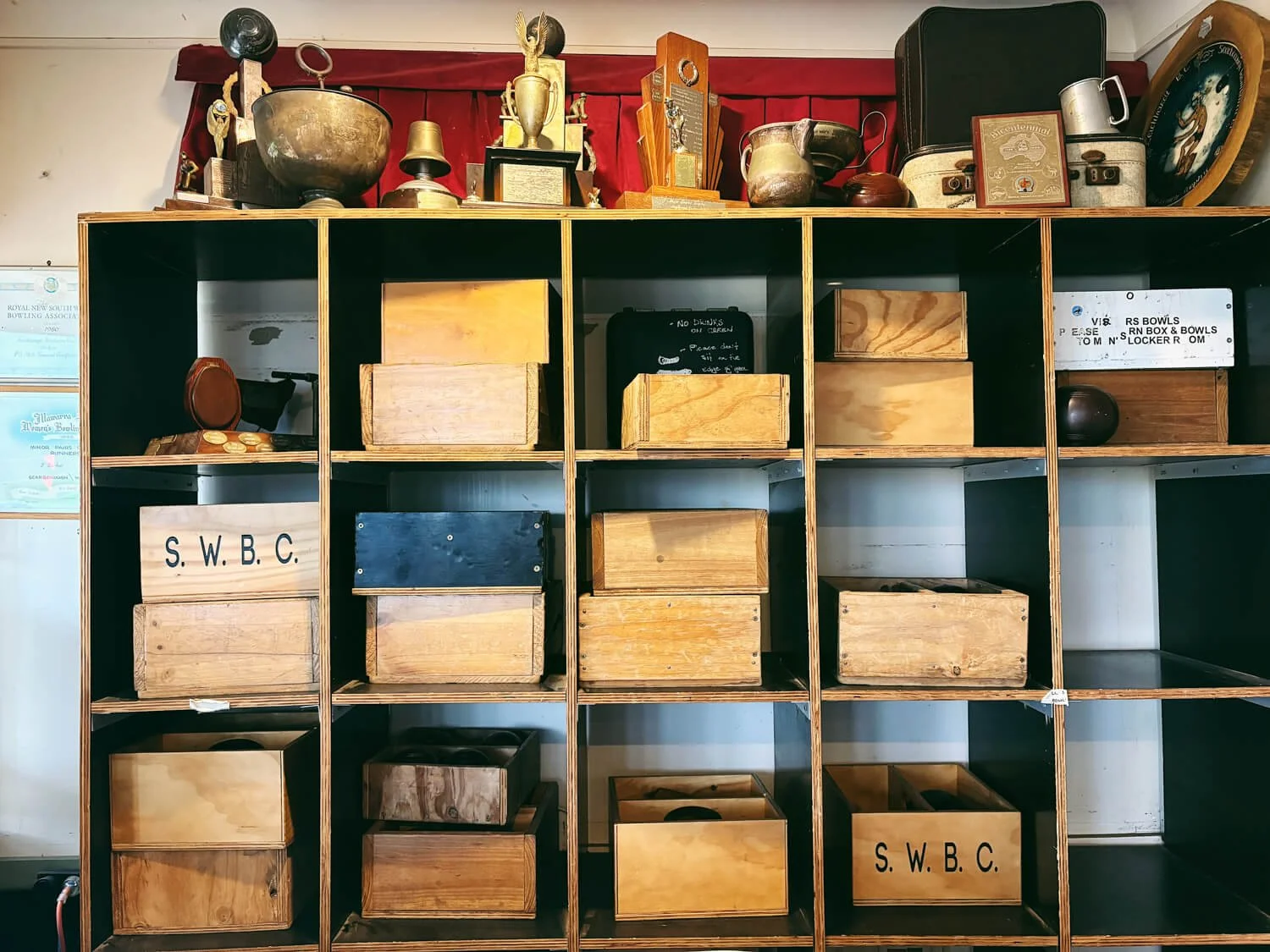 Shelf with wooden boxes and trophies on top, against a white wall with red curtain.