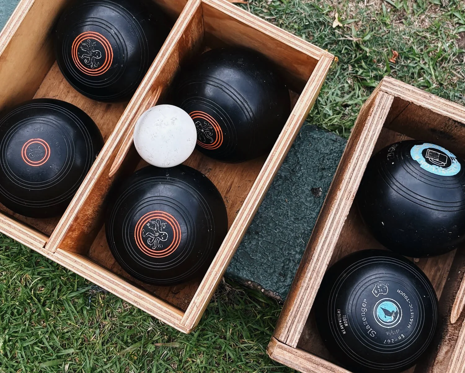 Wooden boxes containing black bocce balls with logos and a white marble, on grass outdoors.