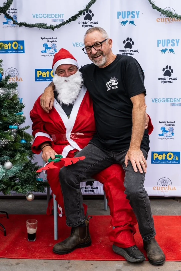 A man dressed as Santa Claus sitting on a chair, holding a Christmas decoration, and a man in a black T-shirt and jeans sitting on Santa's lap. They are both smiling and posing at a festive event with a Christmas tree and a backdrop with various pet-related logos.
