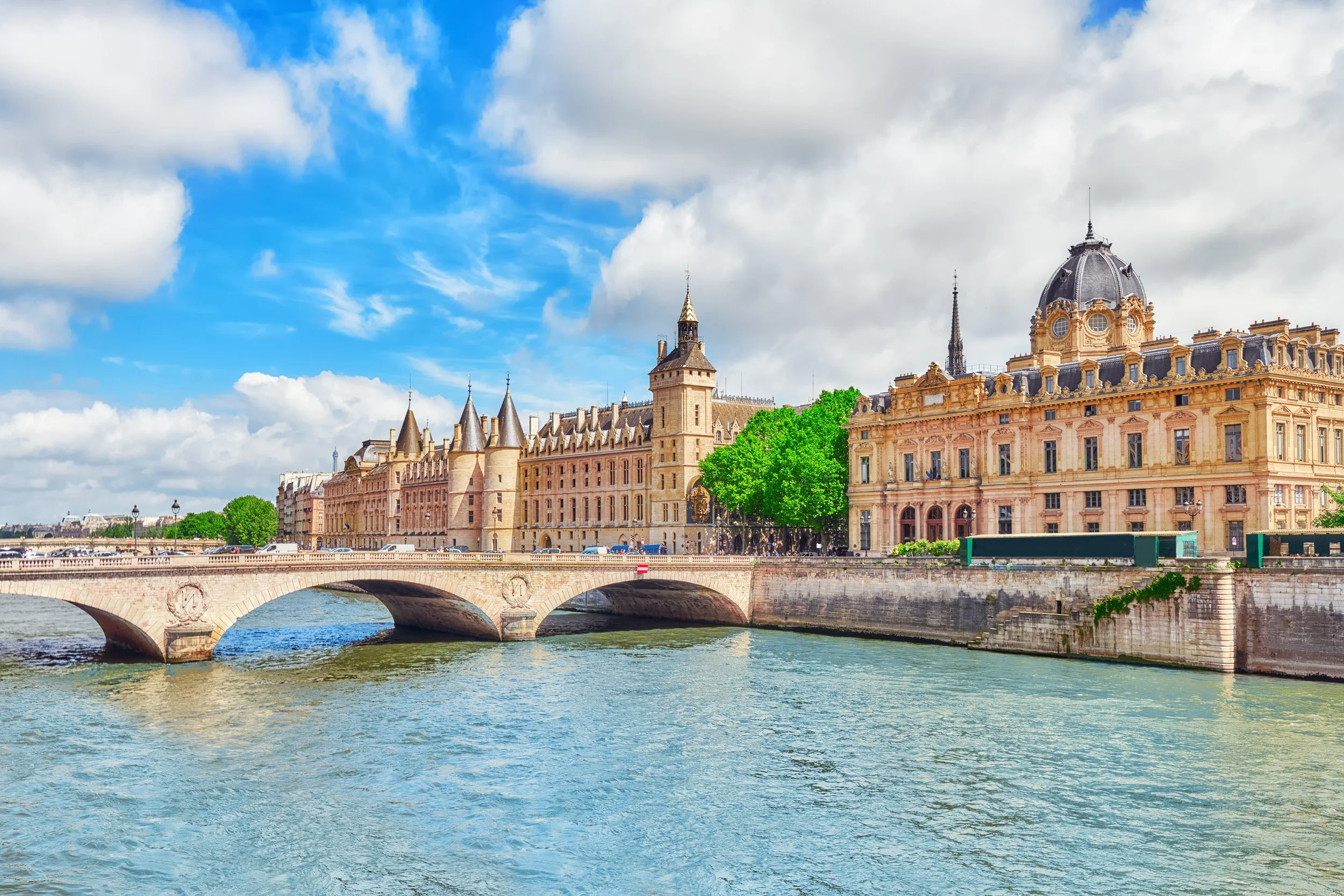 Vue du fleuve Seine avec le pont au premier plan, et des bâtiments historiques, dont le Château de Conciergerie, au fond, sous un ciel partiellement nuageux à Paris.