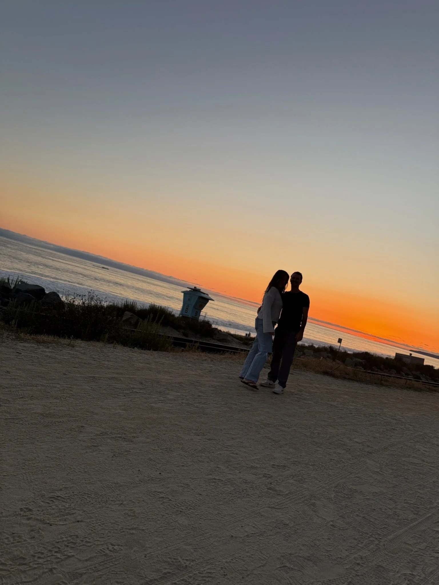 Two people standing close together on a beach at sunset, with the ocean and sky in the background.
