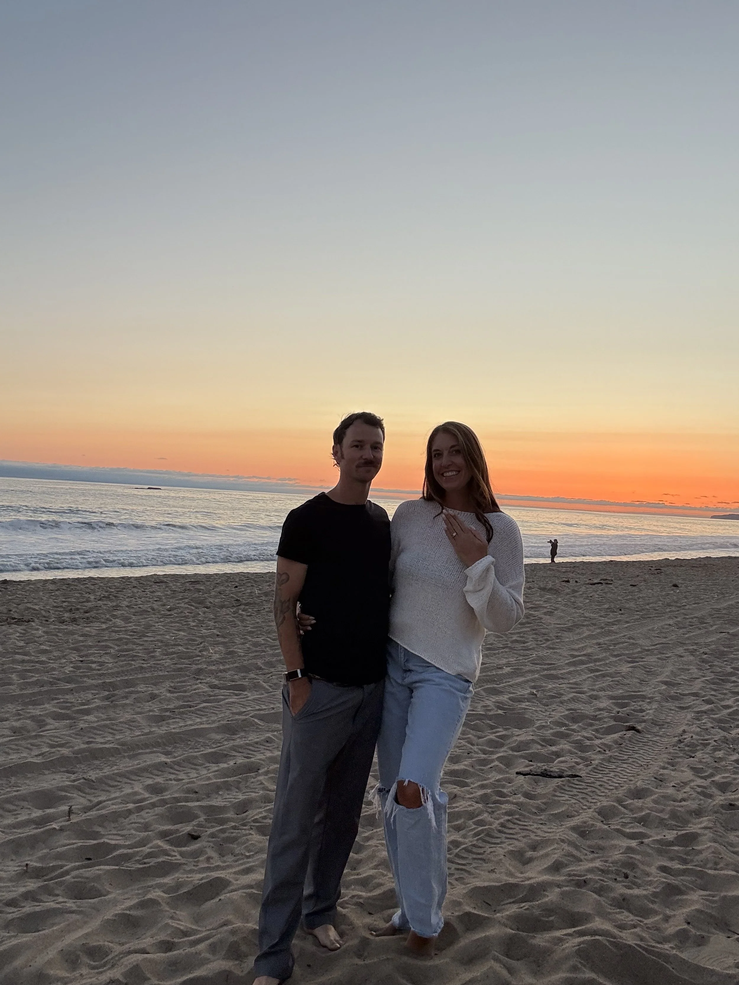 A smiling couple standing on a sandy beach during sunset, with the ocean and colorful sky in the background.