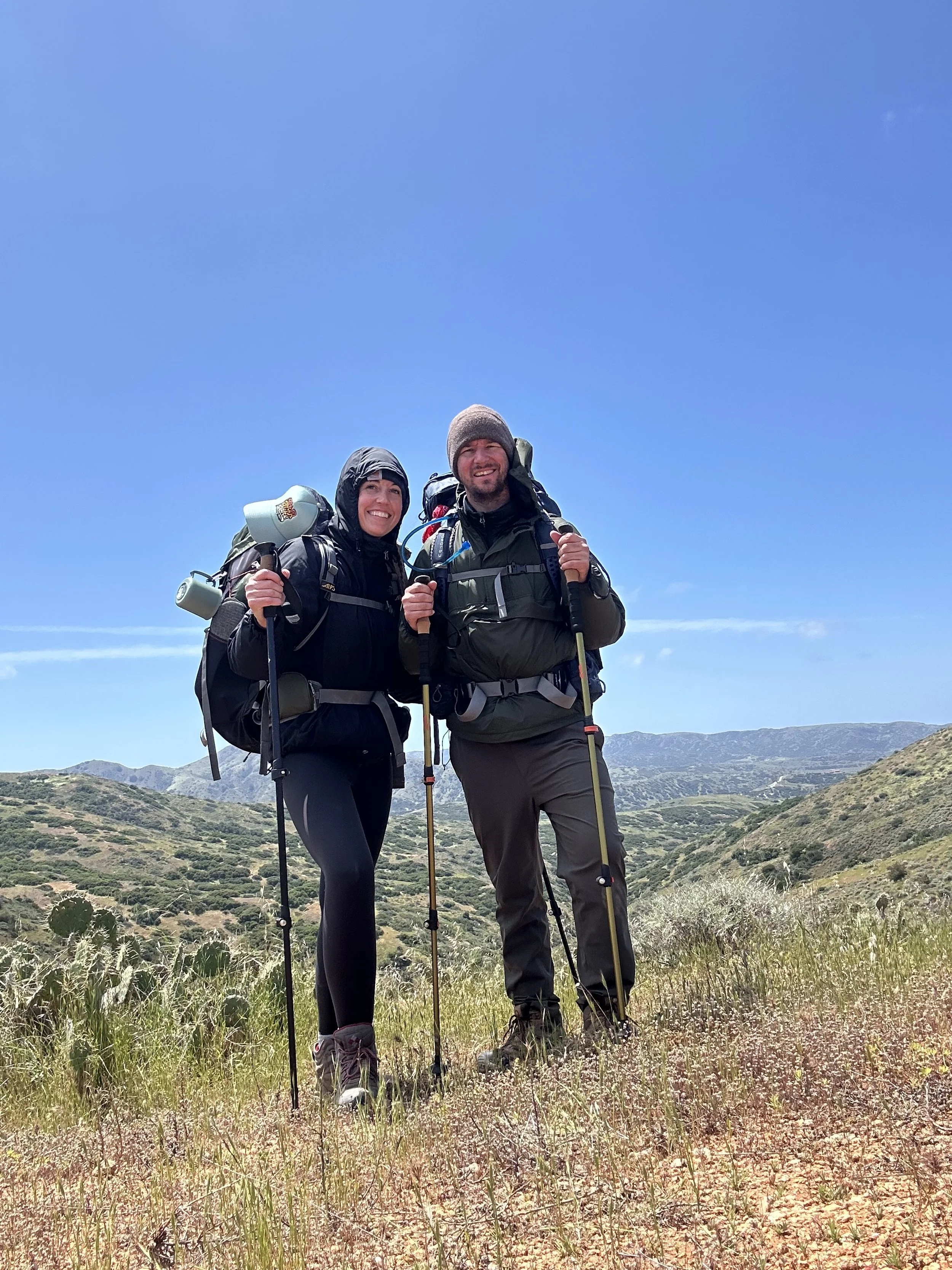 Two hikers with backpacks standing on a grassy trail in a mountainous area under a clear blue sky.