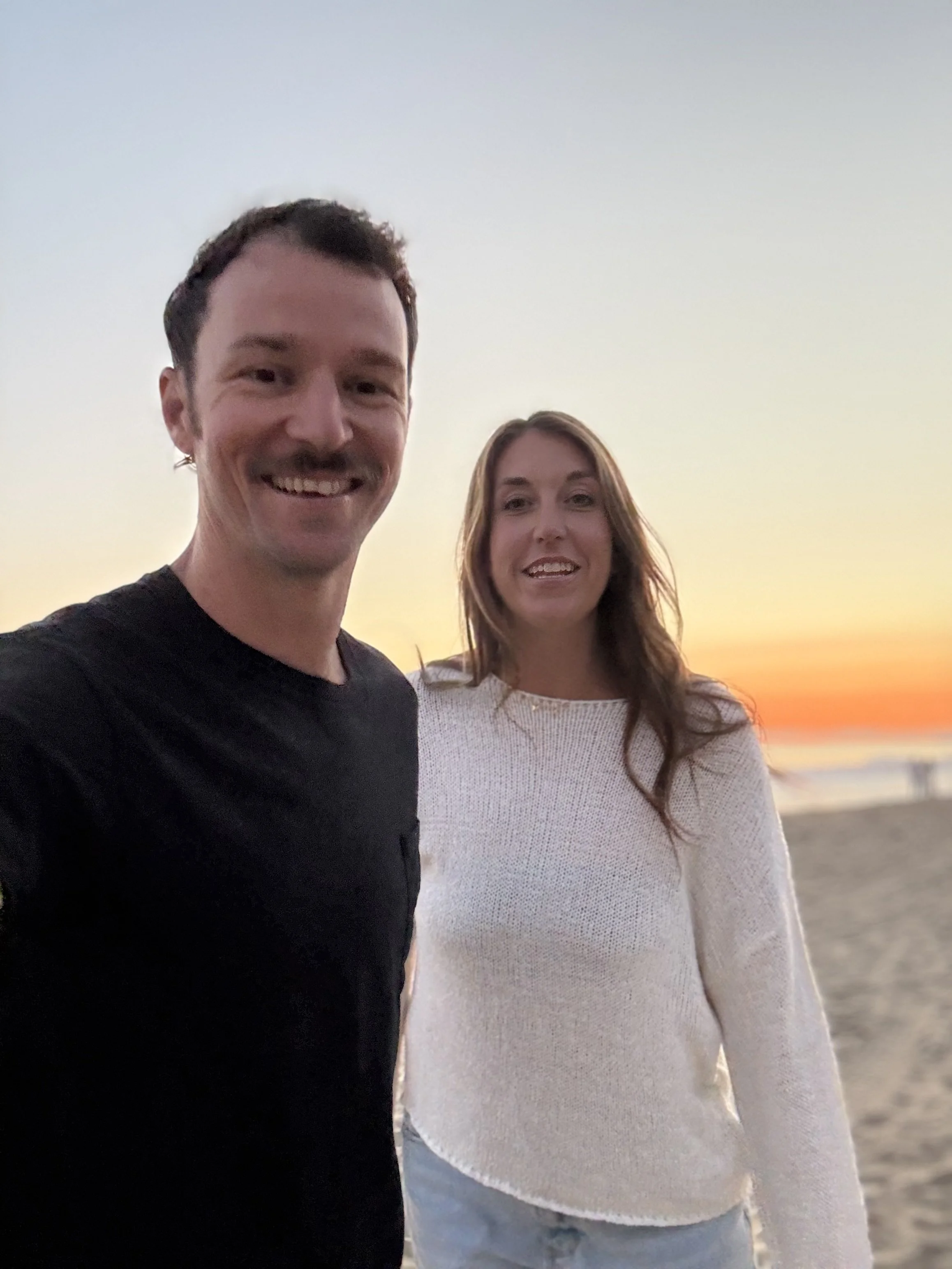 A smiling man and woman taking a selfie on the beach during sunset, with a colorful sky and calm ocean in the background.