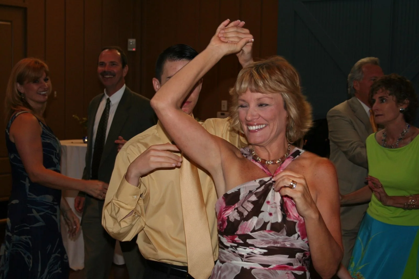 A woman with blonde hair in a floral dress is dancing and smiling with her arm raised at a social event, with other people dancing and talking in the background.