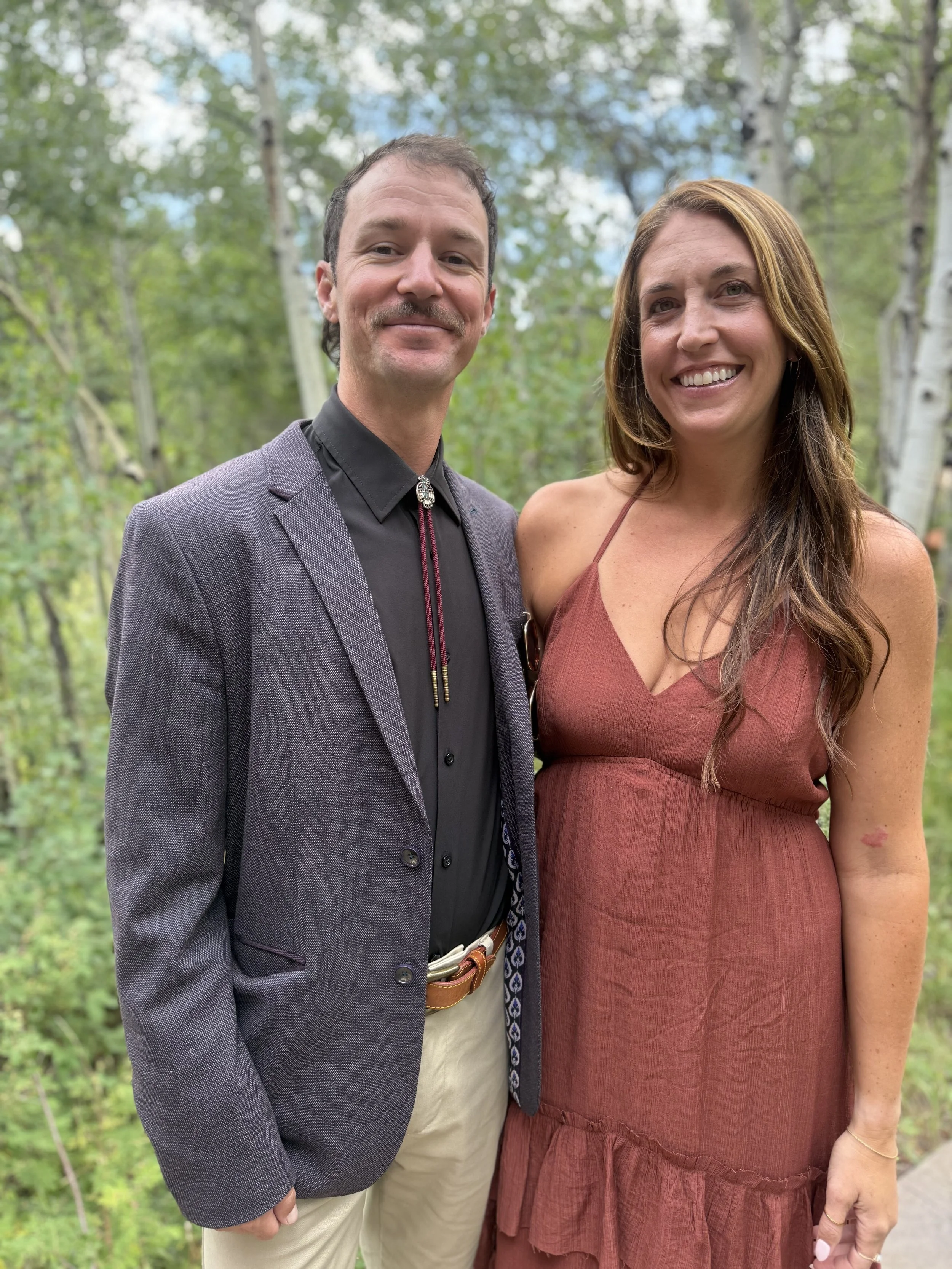 A man and woman smiling outdoors in a forest with green trees in the background. The man is wearing a gray blazer, black shirt, and light-colored pants, while the woman is wearing a brown dress with thin straps.