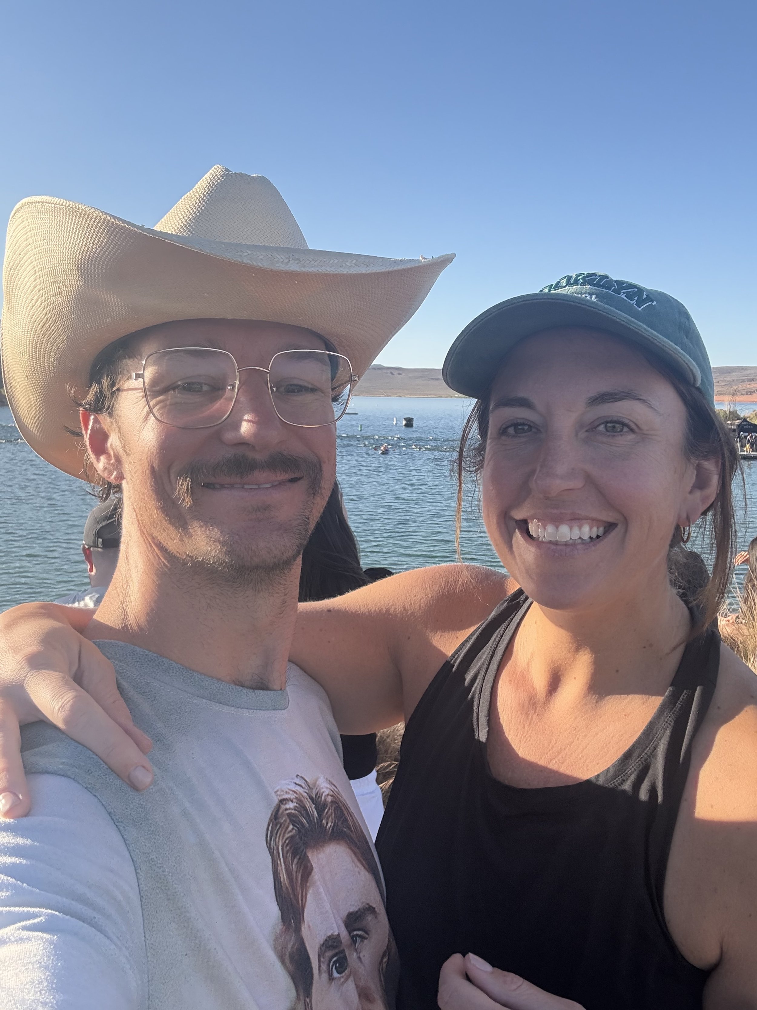A man and woman taking a selfie outdoors near a body of water on a sunny day, both smiling. The man is wearing glasses, a cowboy hat, and a graphic T-shirt. The woman is wearing a dark cap and a black sleeveless top.